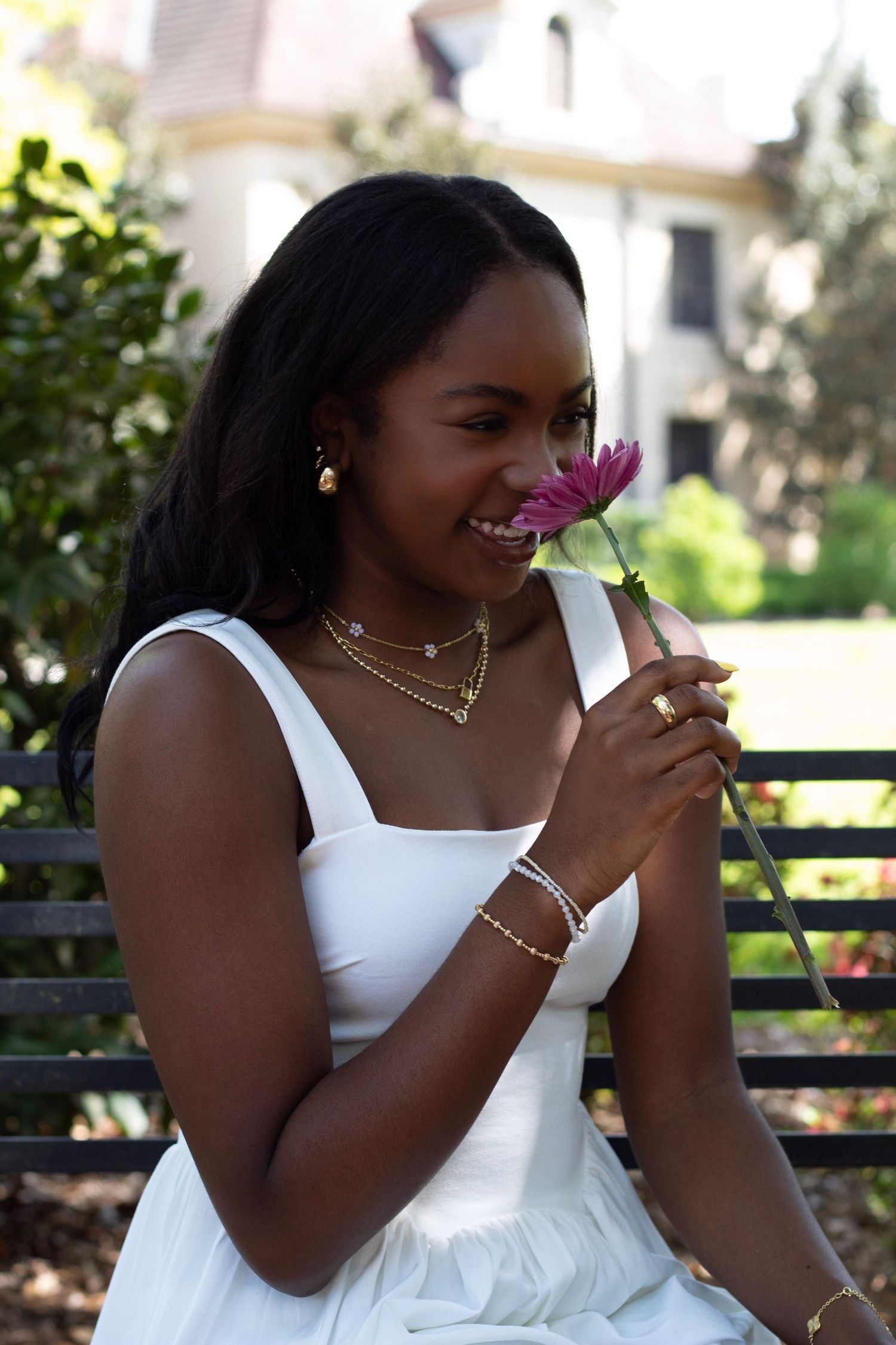 Young Black woman in white dress smelling a pink flower on a bench outdoors.