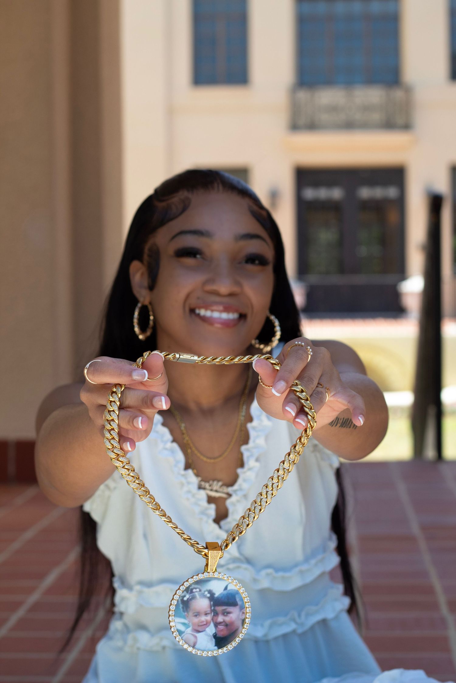 Young woman smiling and holding a gold chain necklace with a photo pendant toward the camera outdoors.