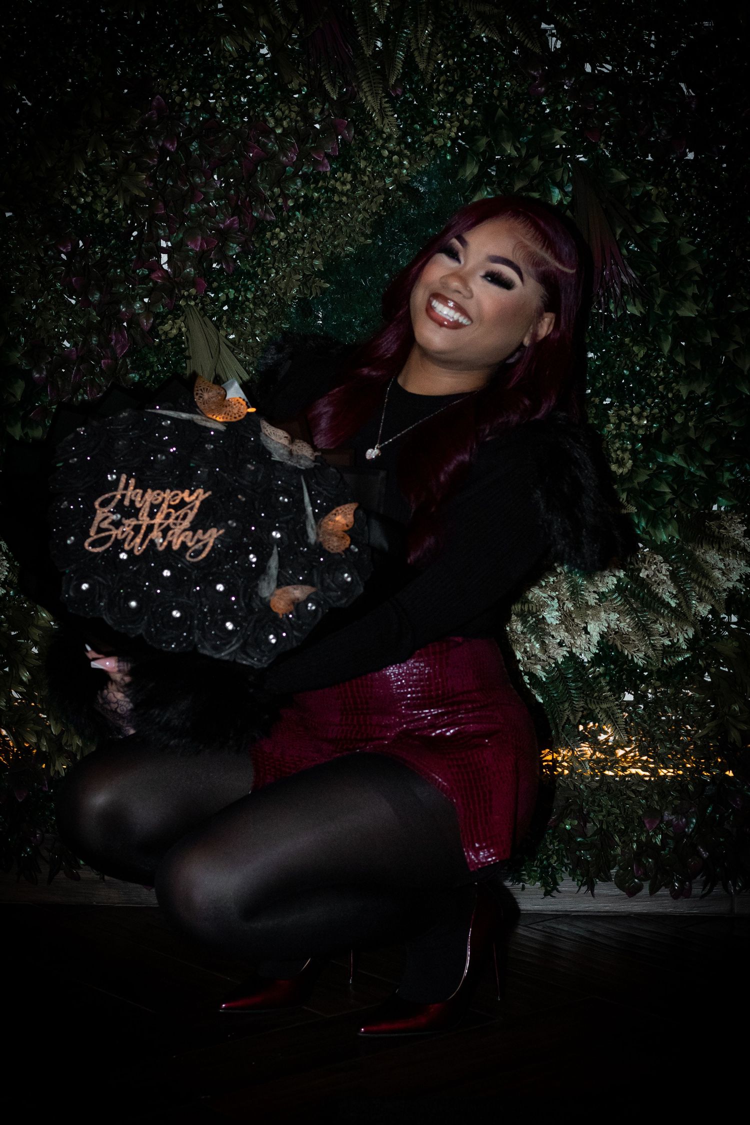 Woman with red hair smiling and holding black Happy Birthday cake against decorative green foliage backdrop.