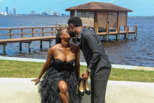 Couple sharing a romantic moment by a waterfront pier on a sunny day in formal attire.