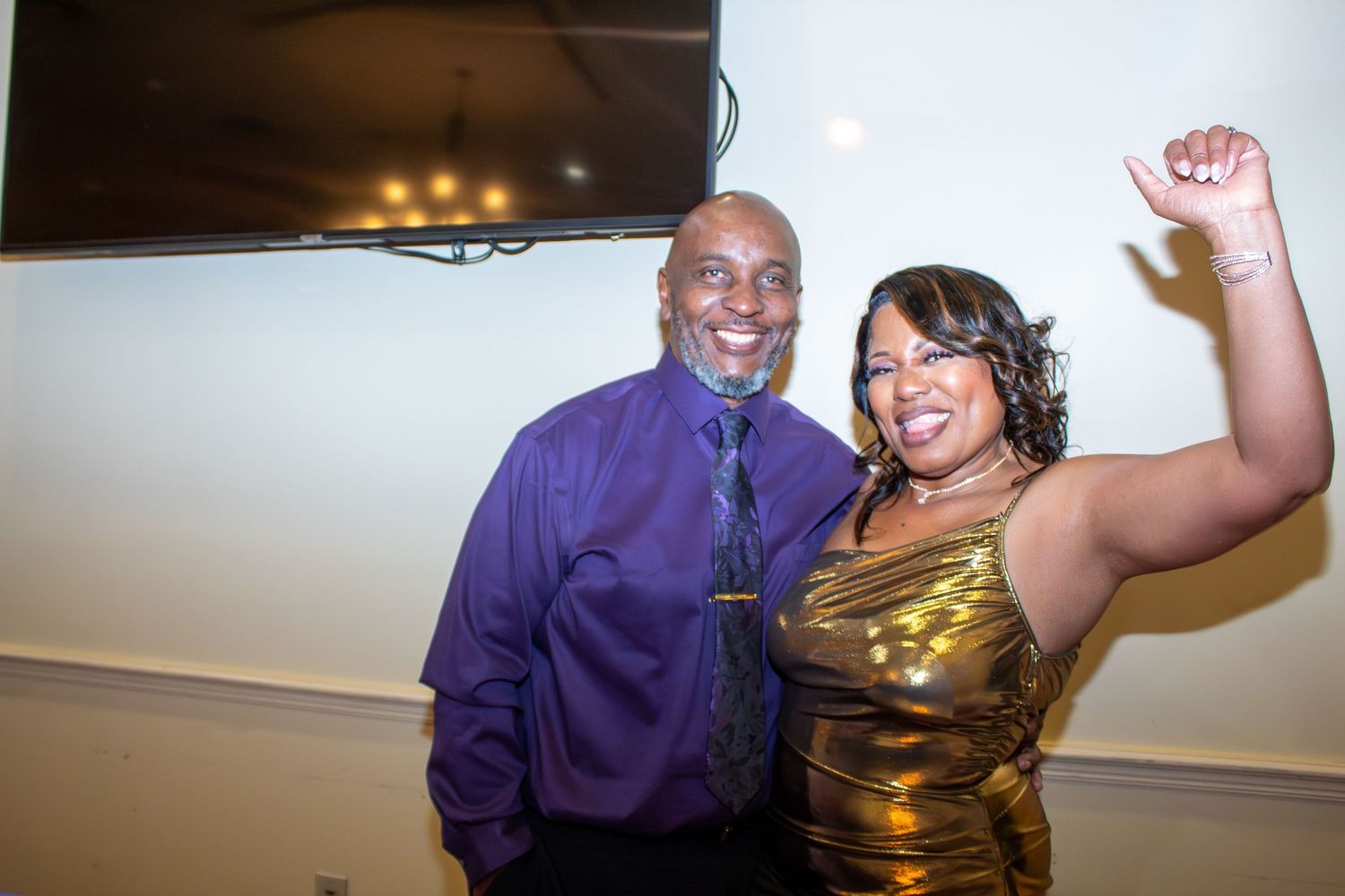 Man in purple shirt and woman in gold dress celebrating joyfully at an indoor event.