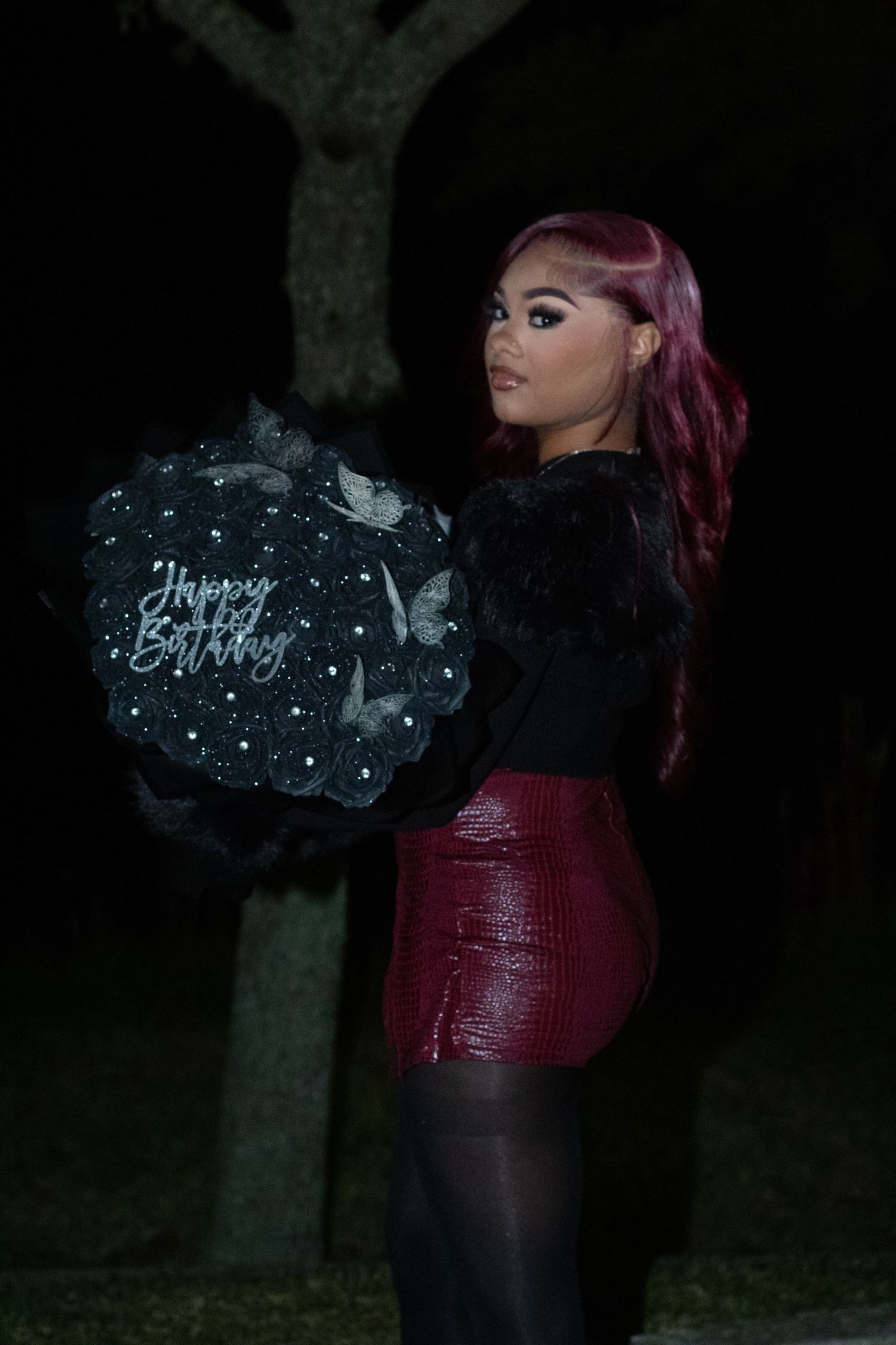 Woman with red hair holding a black Happy Birthday balloon bouquet outdoors at night in a red skirt.