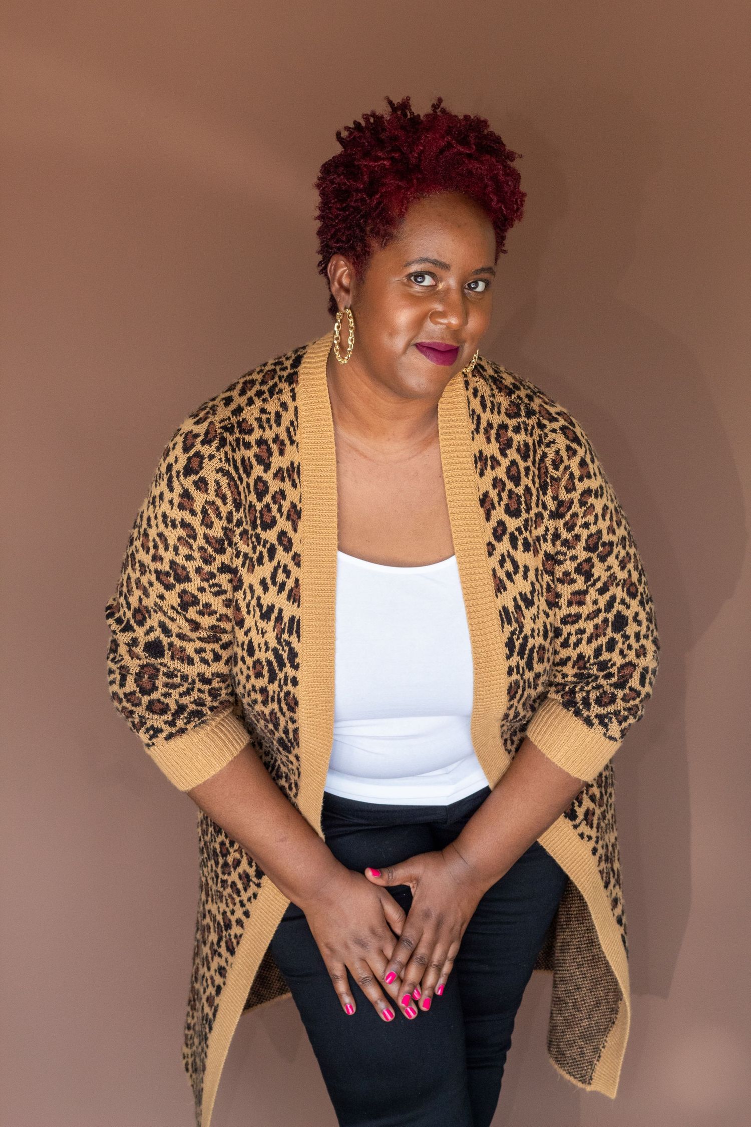Woman with red hair wearing leopard print cardigan posing against brown background.