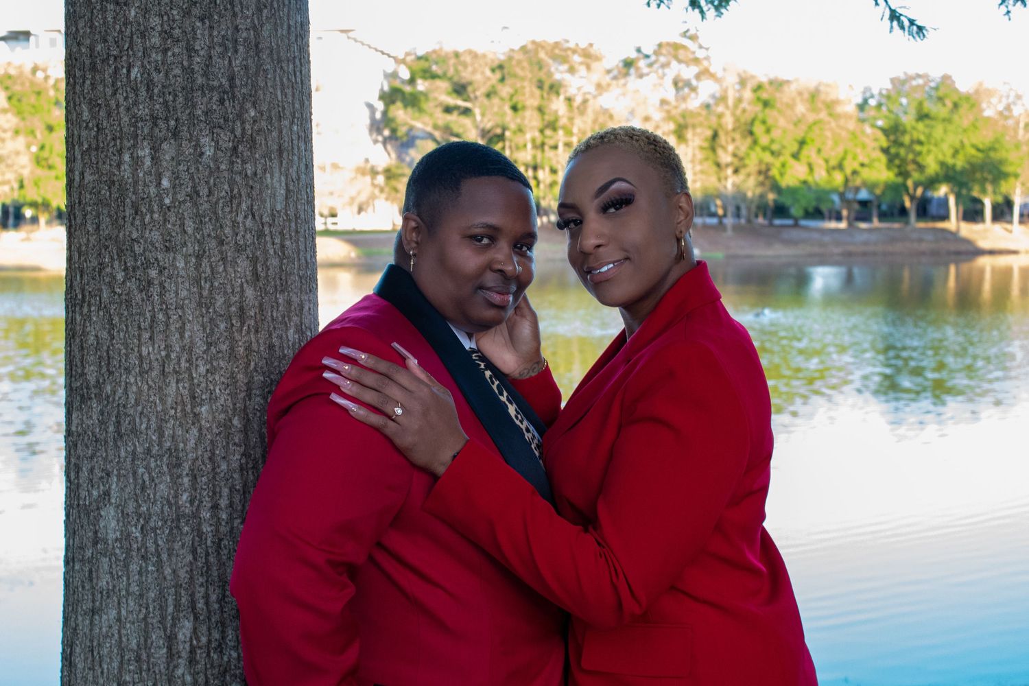 Two women in matching red jackets embracing near a tree by a lake.