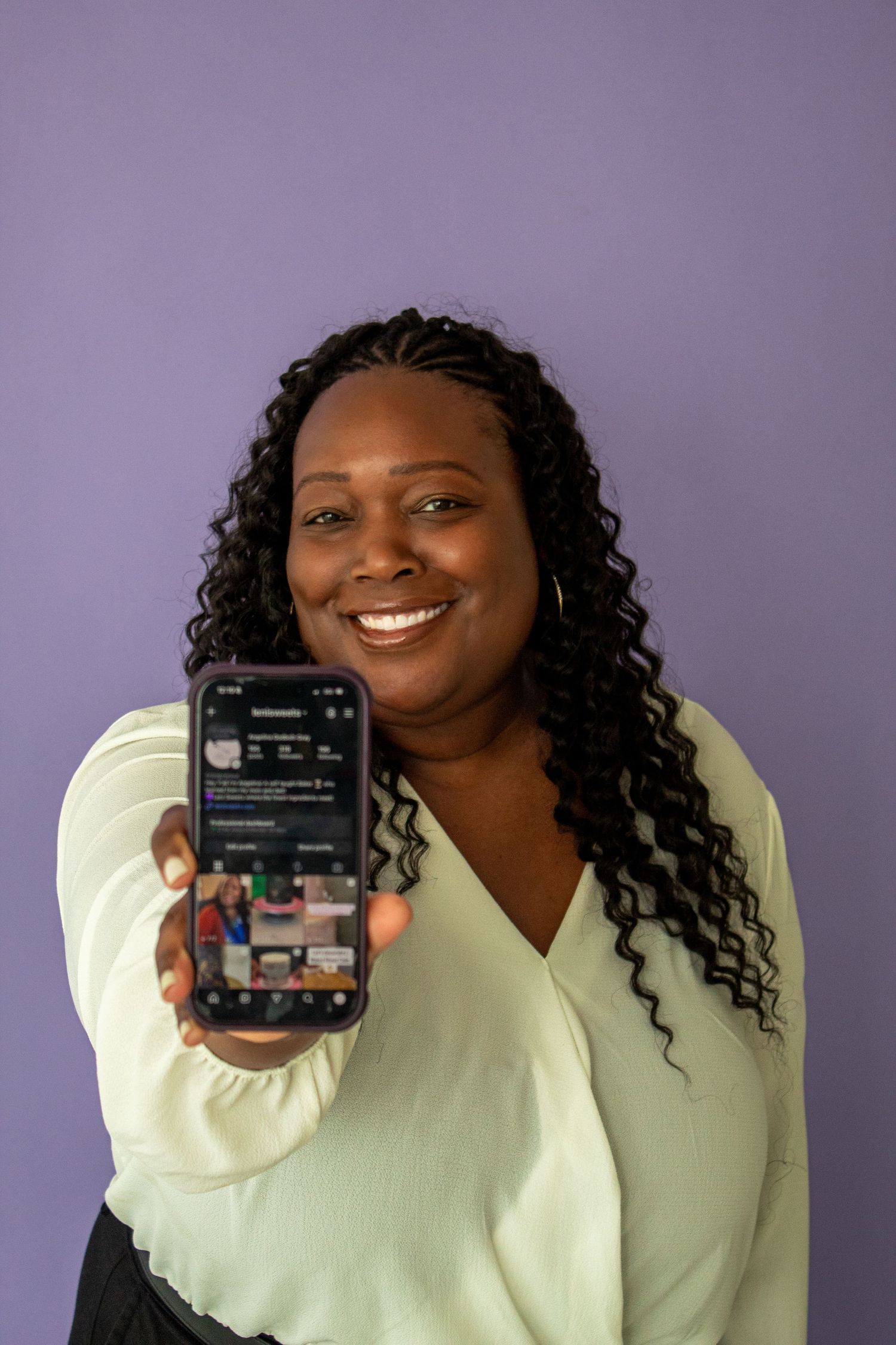 Smiling woman in mint green blouse holding up smartphone showing social media profile against purple background.