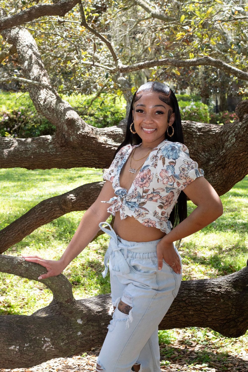 Smiling young woman in floral crop top and jeans posing by a tree outdoors.