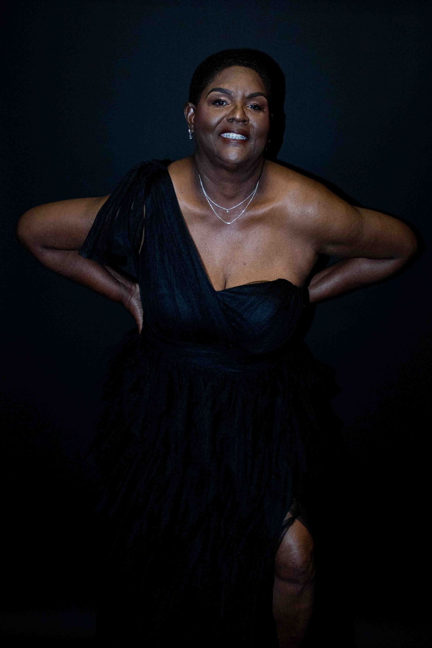 Woman in elegant black one-shoulder dress posing confidently against dark background.