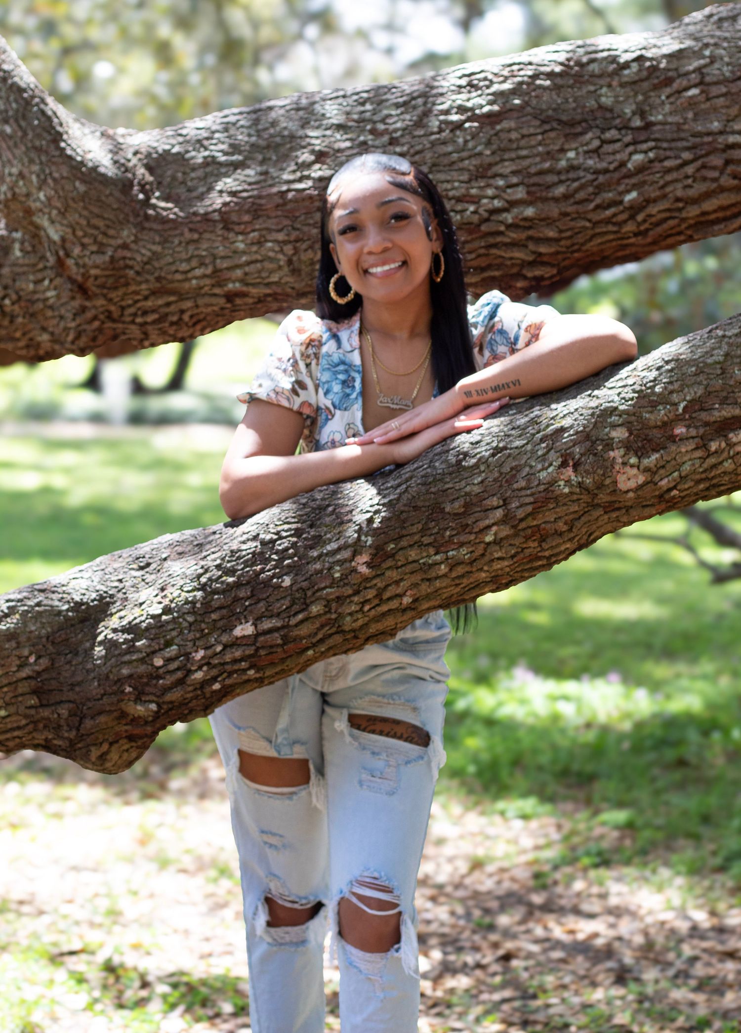 Smiling young woman posing between large tree branches in sunny park wearing floral top and ripped jeans.