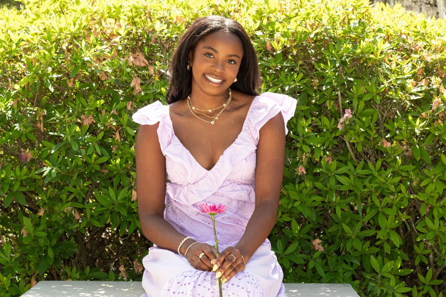 Smiling young woman in a pink floral dress holding a pink flower, seated outdoors near green hedges.