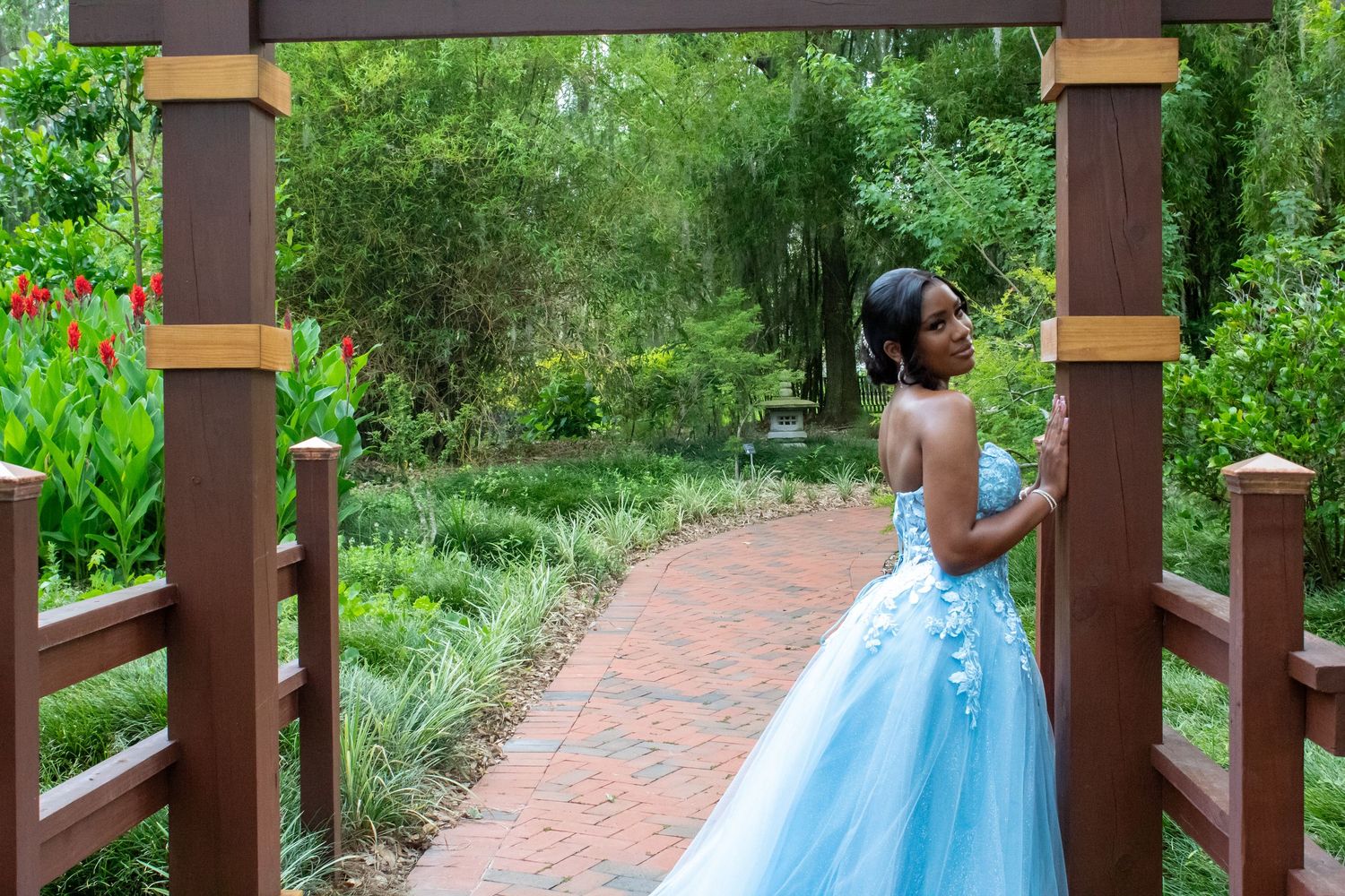 Young girl in blue ball gown stands in garden archway on brick path.