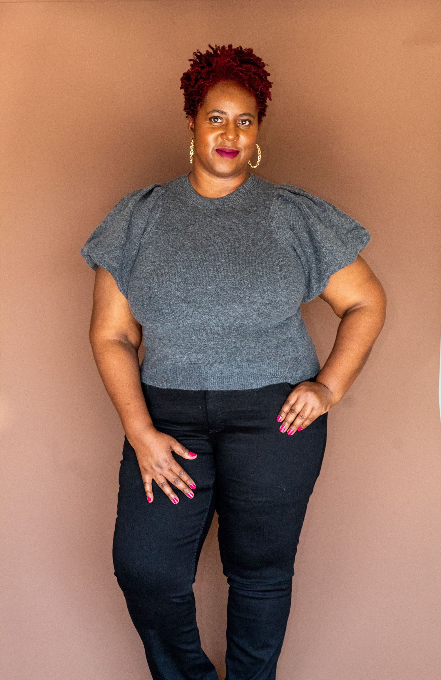 Woman with red hair in gray puff-sleeve top and black jeans posing confidently.