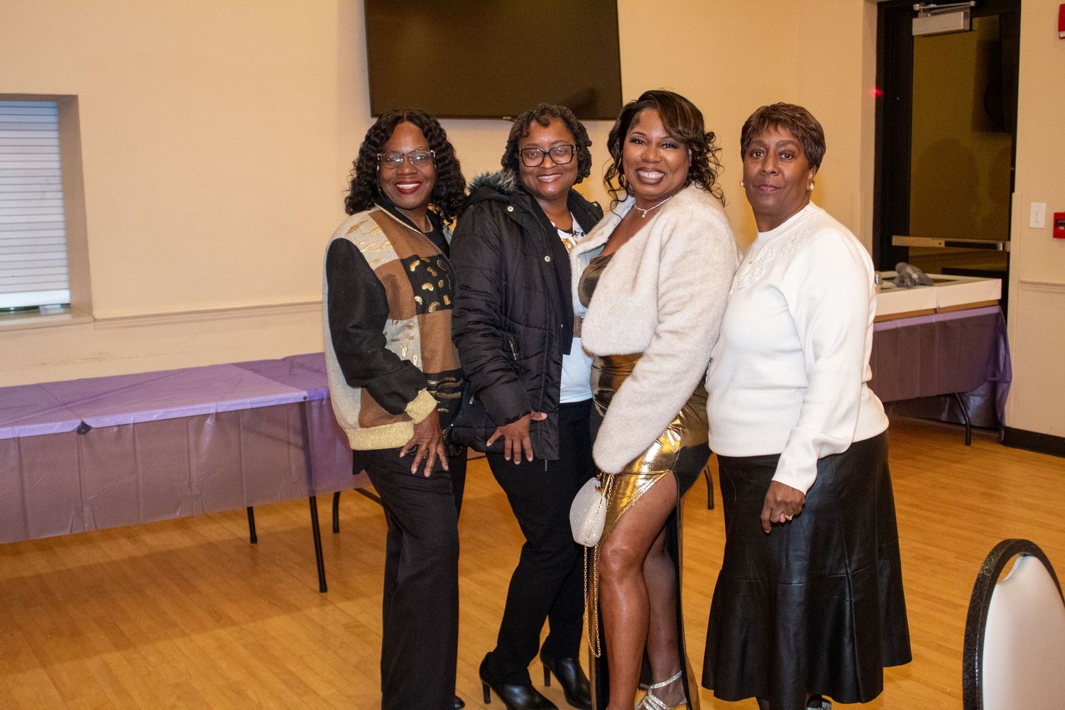 Four women posing together and smiling at an indoor event with purple table decorations.