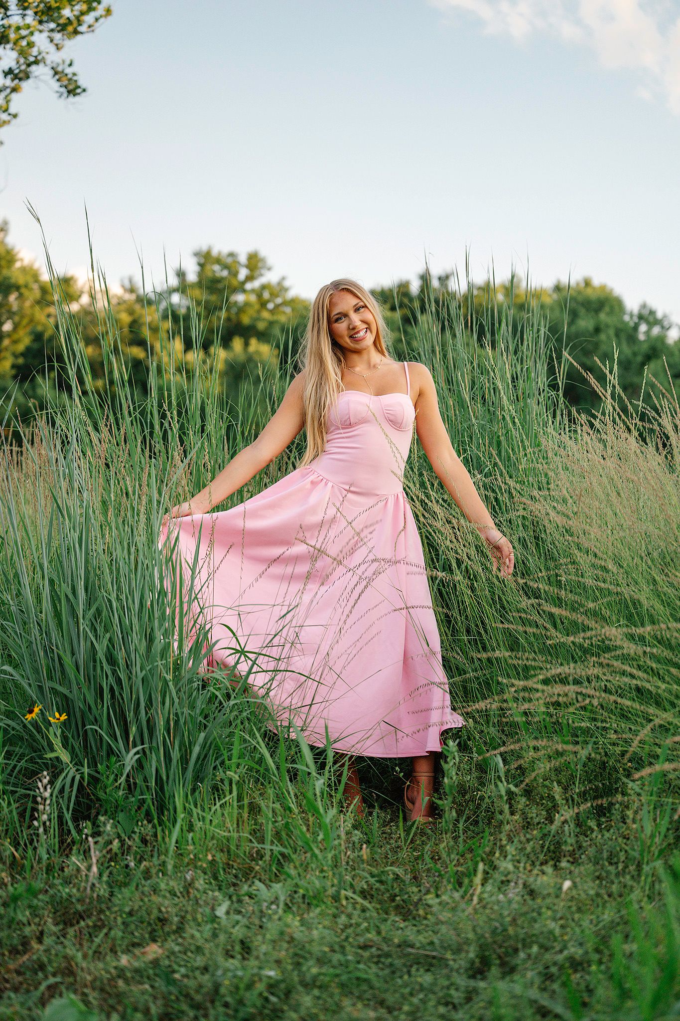 Smiling blonde woman in a flowing pink sundress standing in tall green grass field on a sunny day posing for Ankeny senior photographer