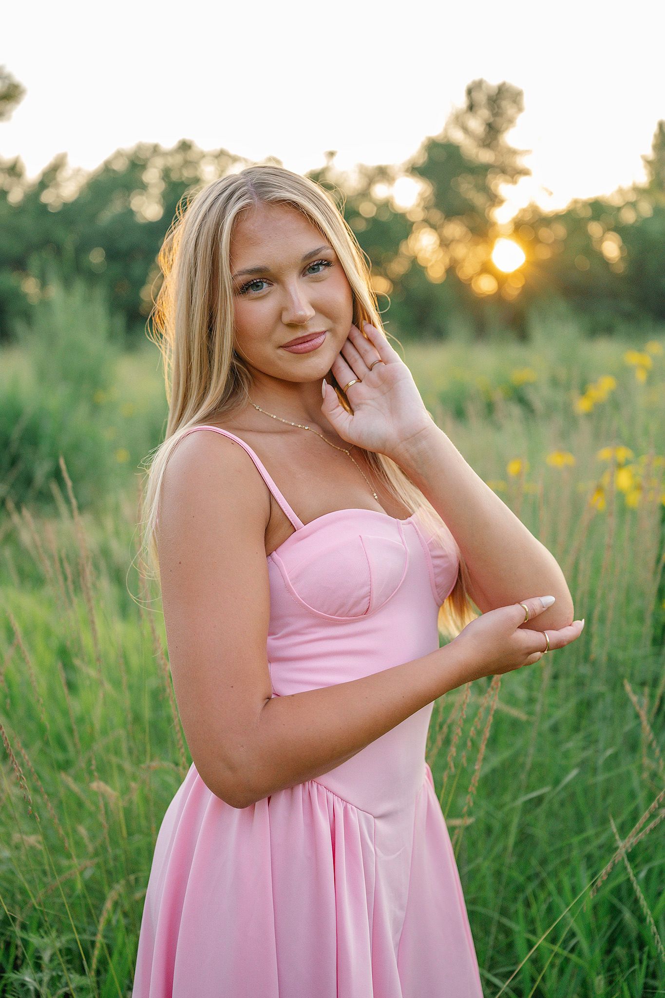Young blonde woman in pink satin dress poses in green meadow at golden sunset.