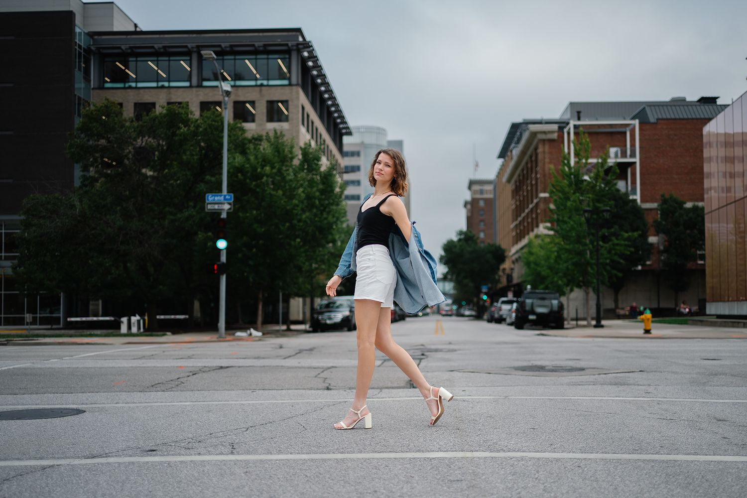 Young woman in white shorts and black top walks across urban street intersection on a cloudy day.