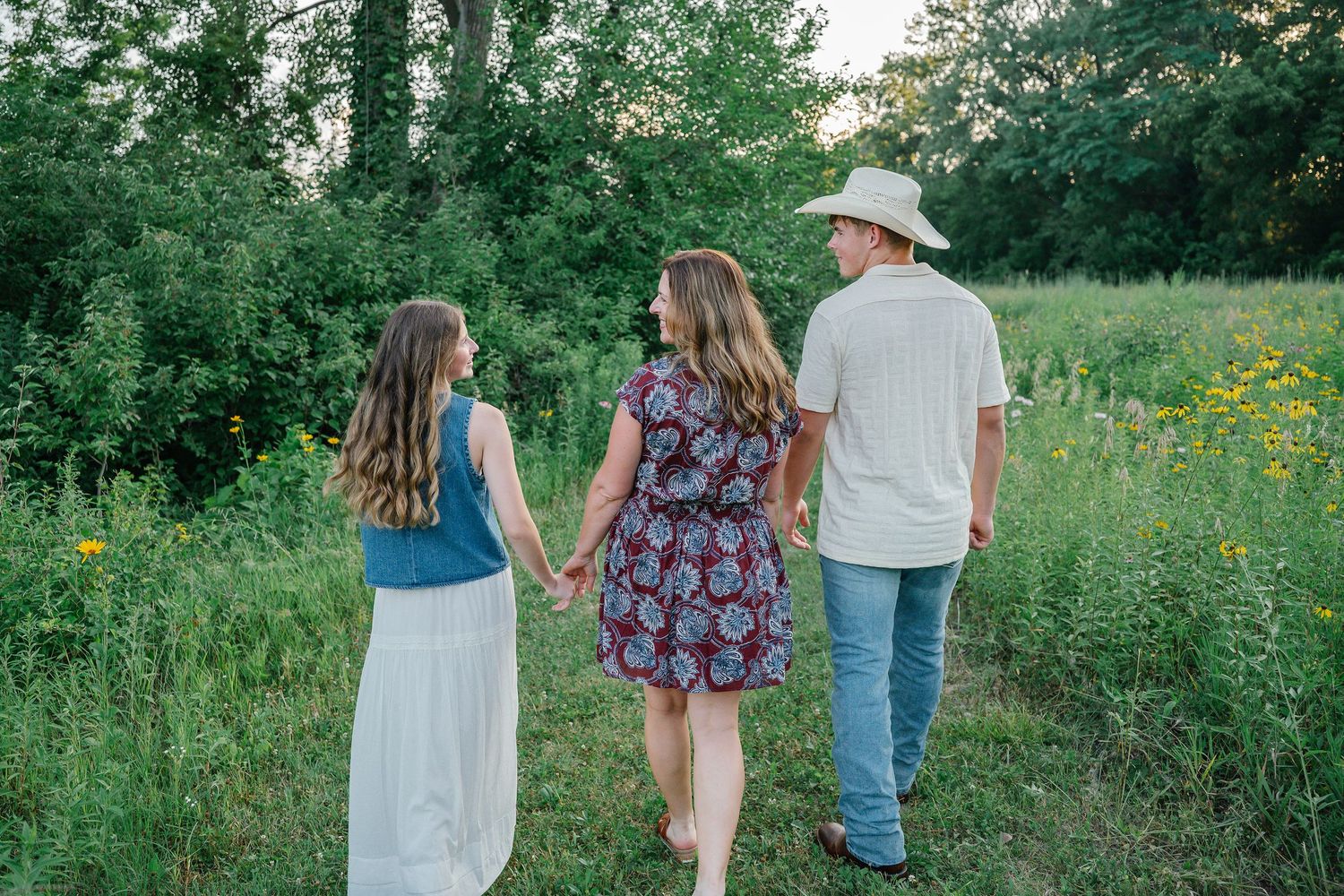 Family of three walking hand-in-hand through a lush green meadow at golden hour, viewed from behind.