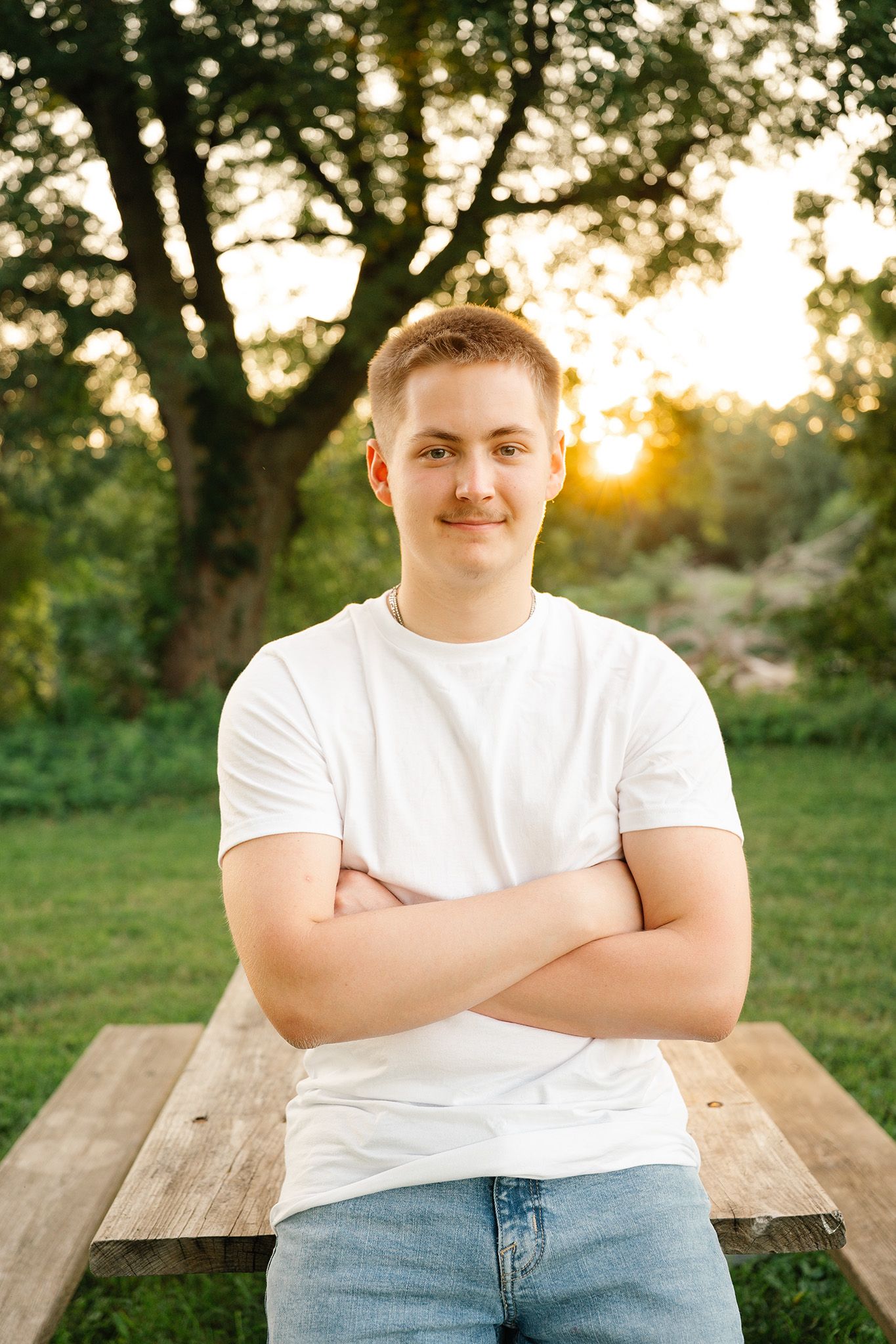 Young man in white t-shirt and jeans sitting at wooden picnic table outdoors at golden hour sunset.