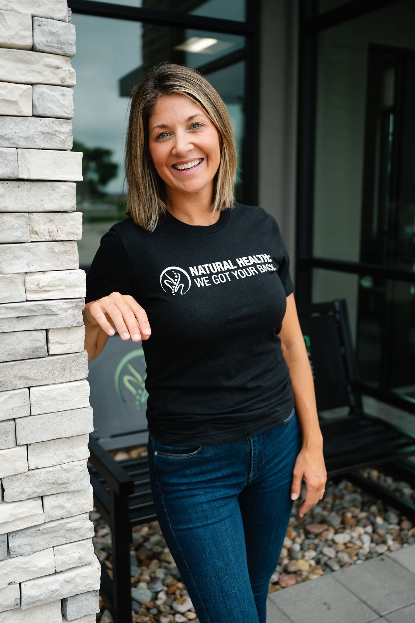 Smiling woman in black Natural Healing t-shirt leaning against stone pillar outside a modern building.