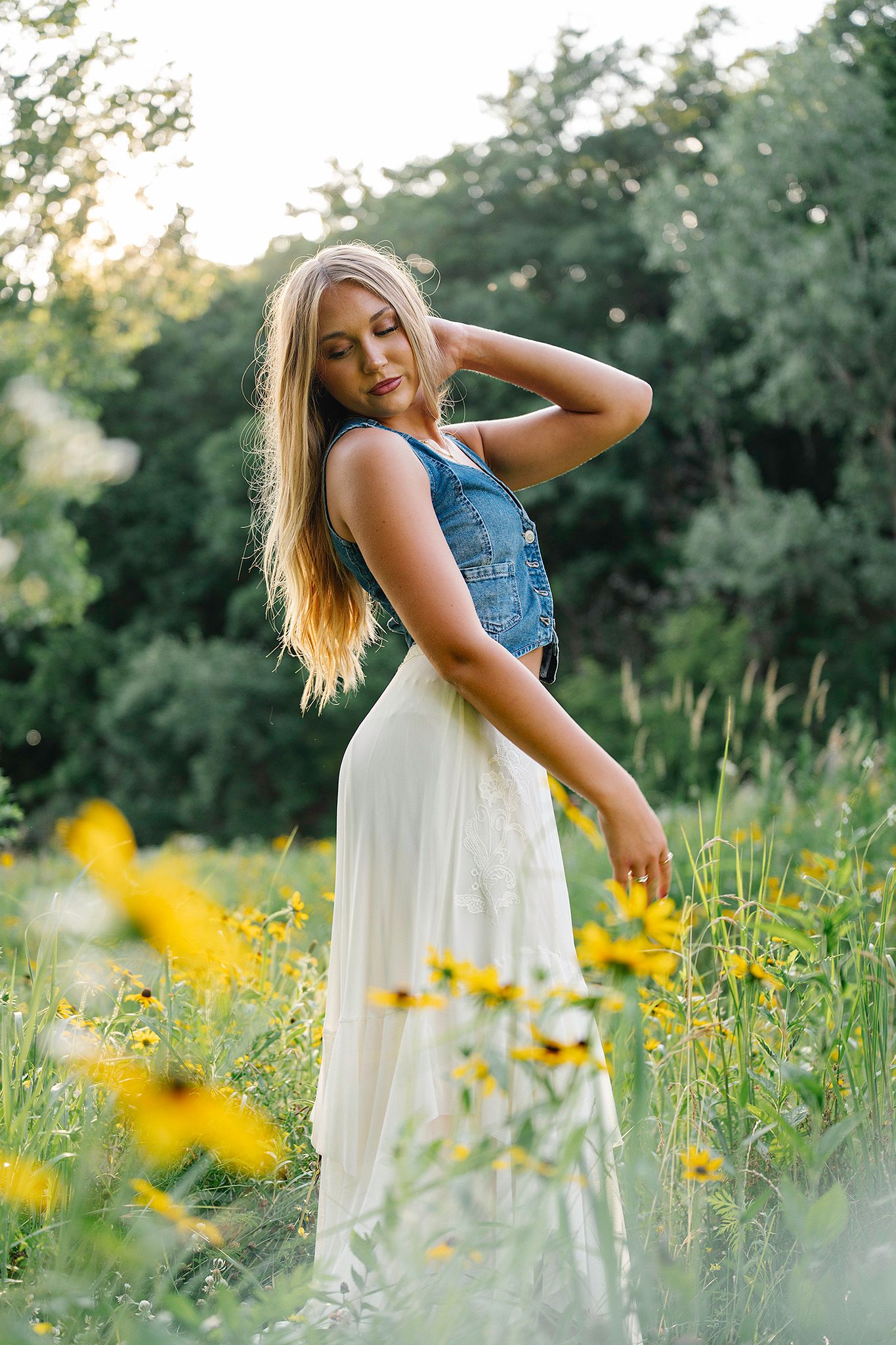 Young blonde woman in denim vest and white skirt posing among yellow wildflowers in a lush green field at sunset.