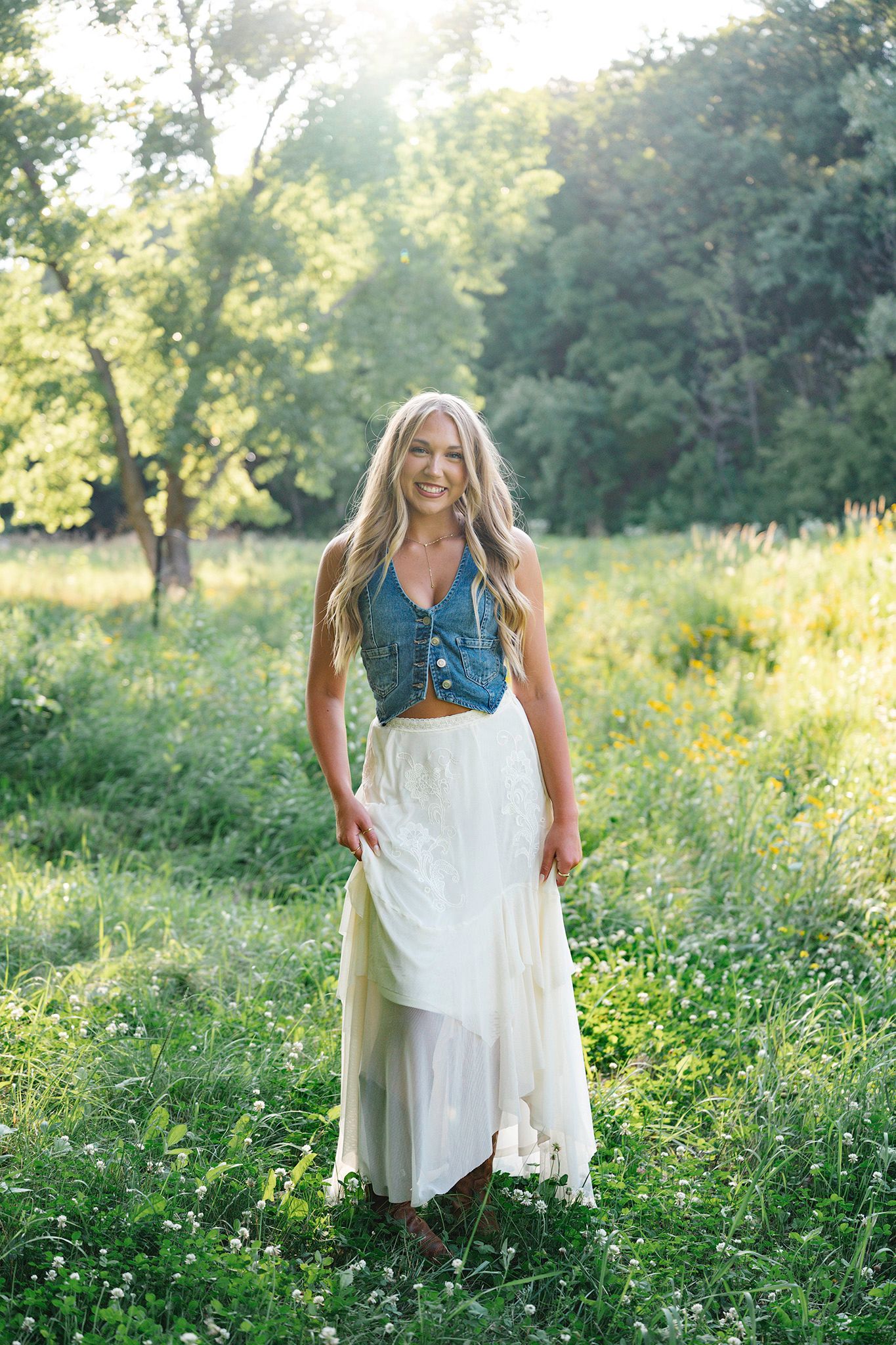 Smiling blonde woman in denim top and maxi skirt standing in a sunlit green meadow for Images by Merron, Ankeny & Des Moines senior photos
