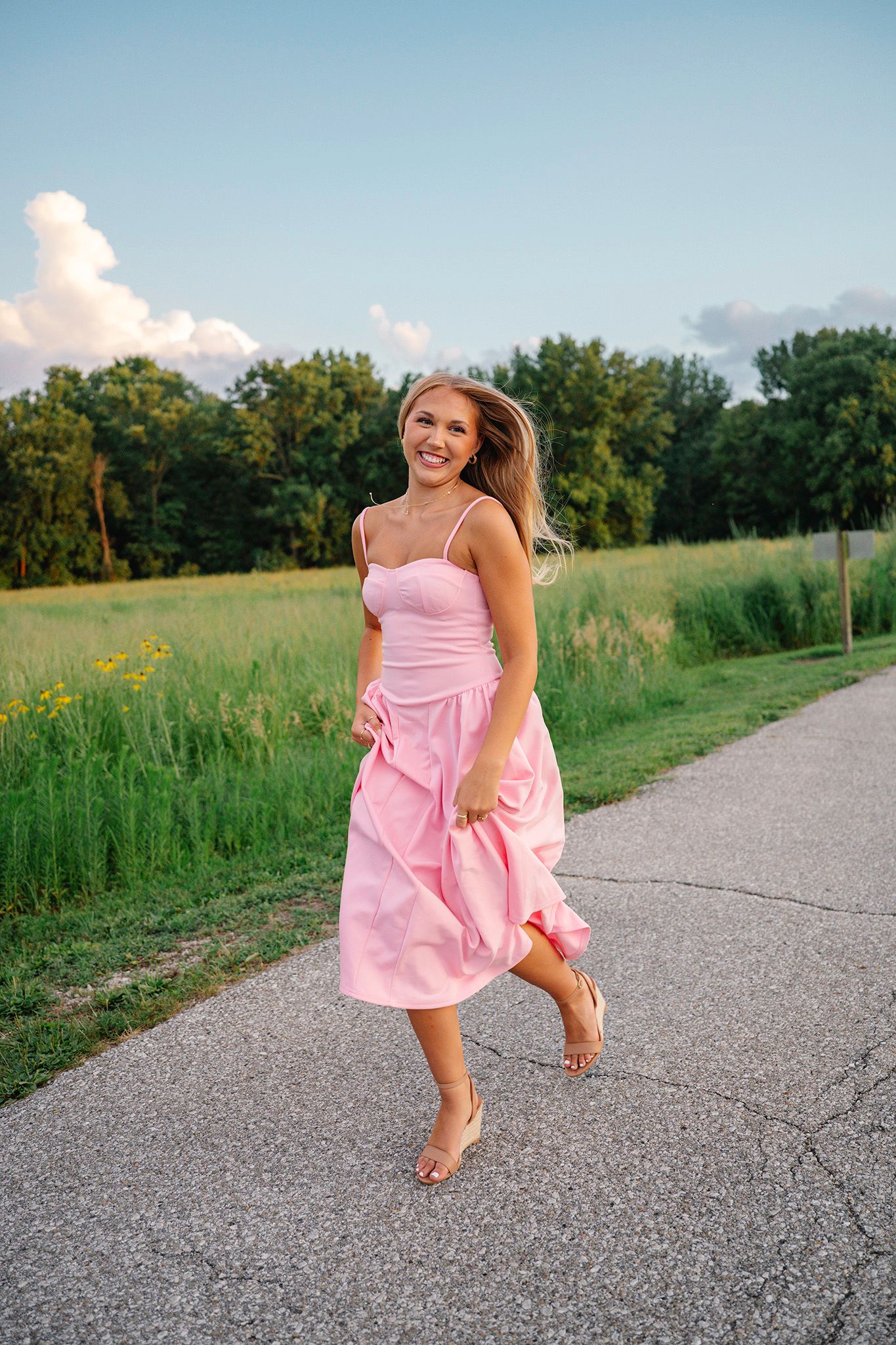 Smiling blonde woman in a pink midi dress walking barefoot on a path surrounded by green fields posing for Ankeny senior photographer