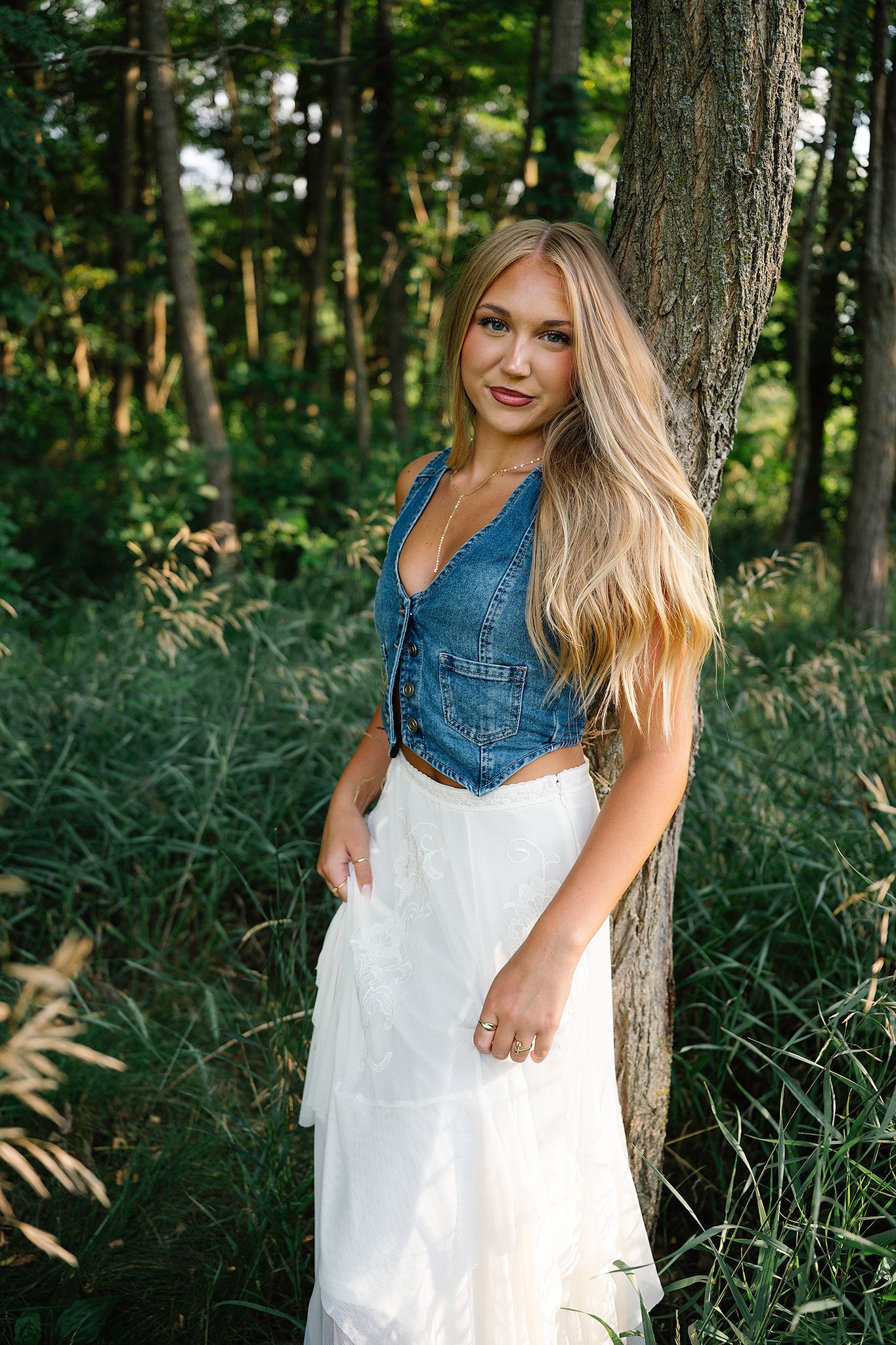 Blonde woman in denim vest and white skirt posing by a tree for Images by Merron, Ankeny & Des Moines senior photographer