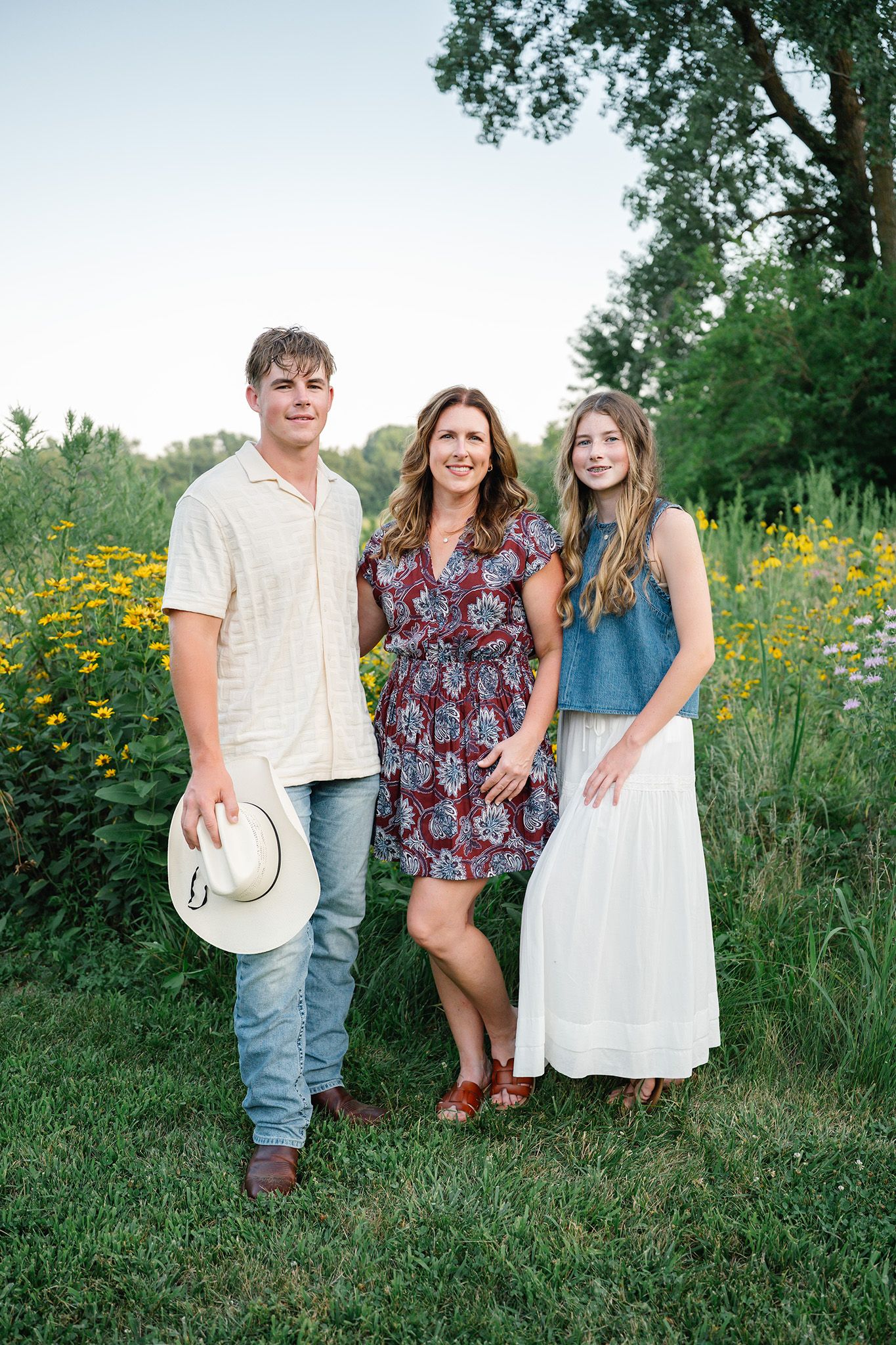A woman and two teens pose together in nature with yellow wildflowers during a family photo session posing for Ankeny family photographer.