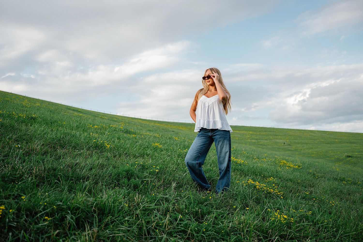 Woman in white top and wide-leg jeans posing in a lush green field with yellow wildflowers under a cloudy sky for Ankeny senior photographer