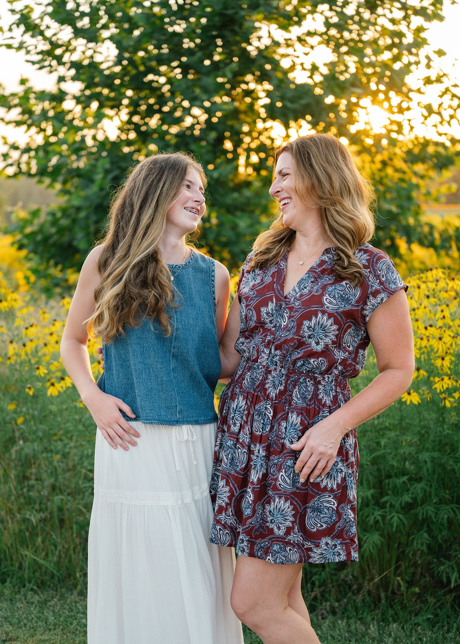 Mother and daughter smiling in floral and denim outfits during golden hour outdoors posing for Ankeny family photographer.