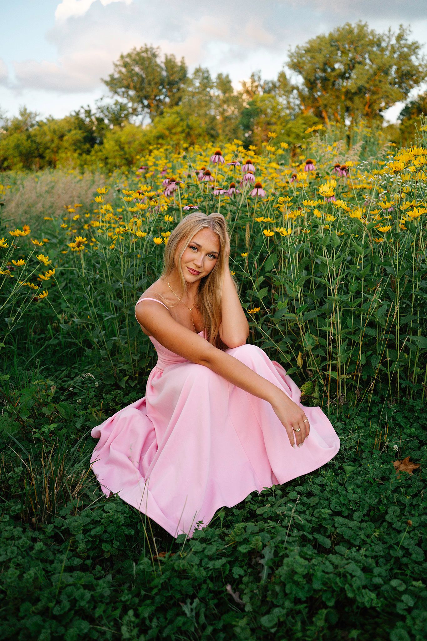 Woman in pink dress sitting among wildflowers in a lush green garden at sunset posing for Ankeny senior photographer