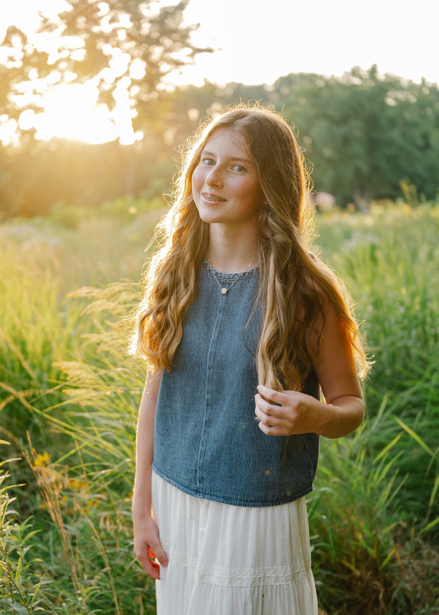 Young girl with long wavy hair in denim top and white skirt smiling in golden sunset field posing for Ankeny family photographer.