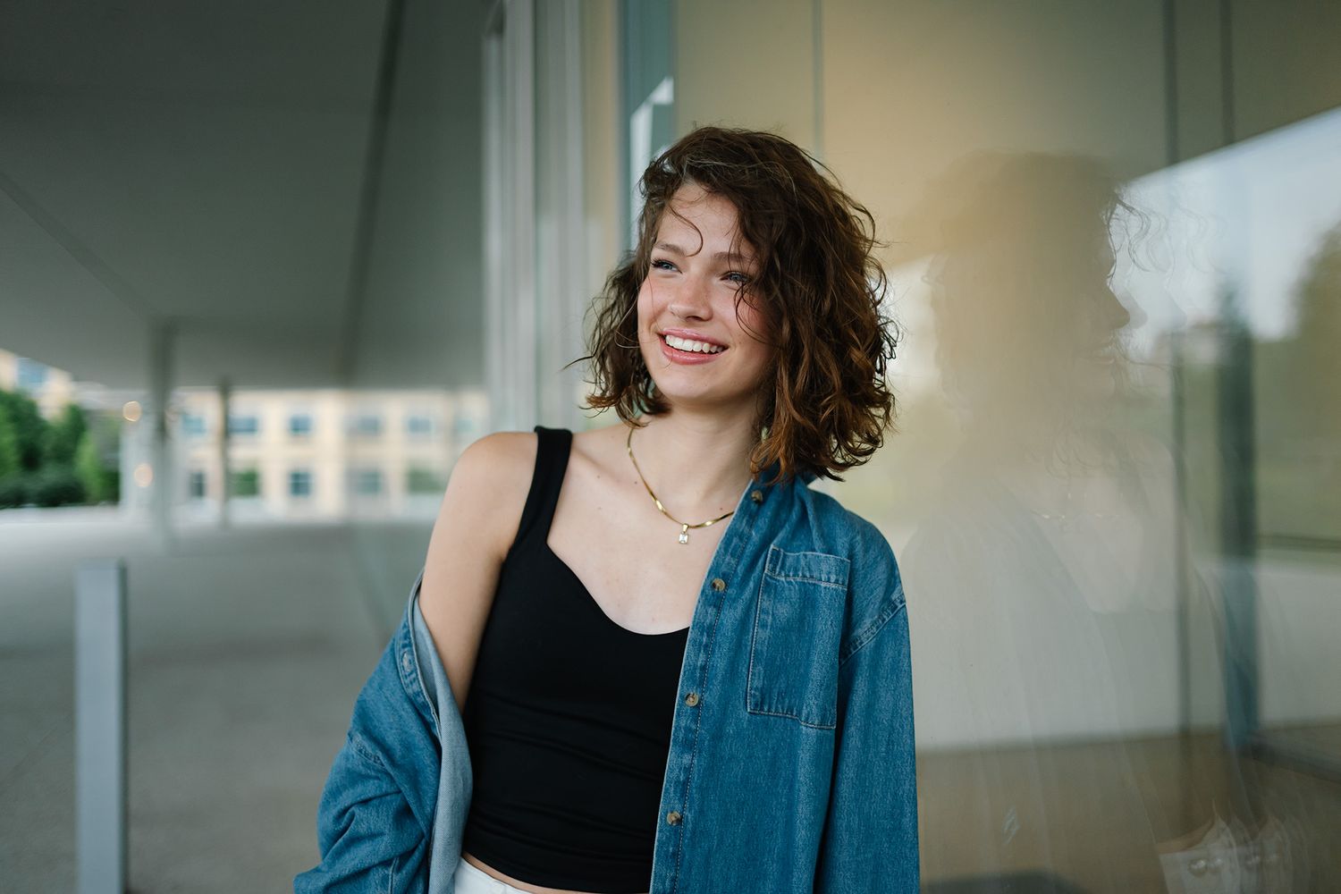 Smiling young woman with curly hair wearing a denim jacket and black top, posing outdoors near a modern building.