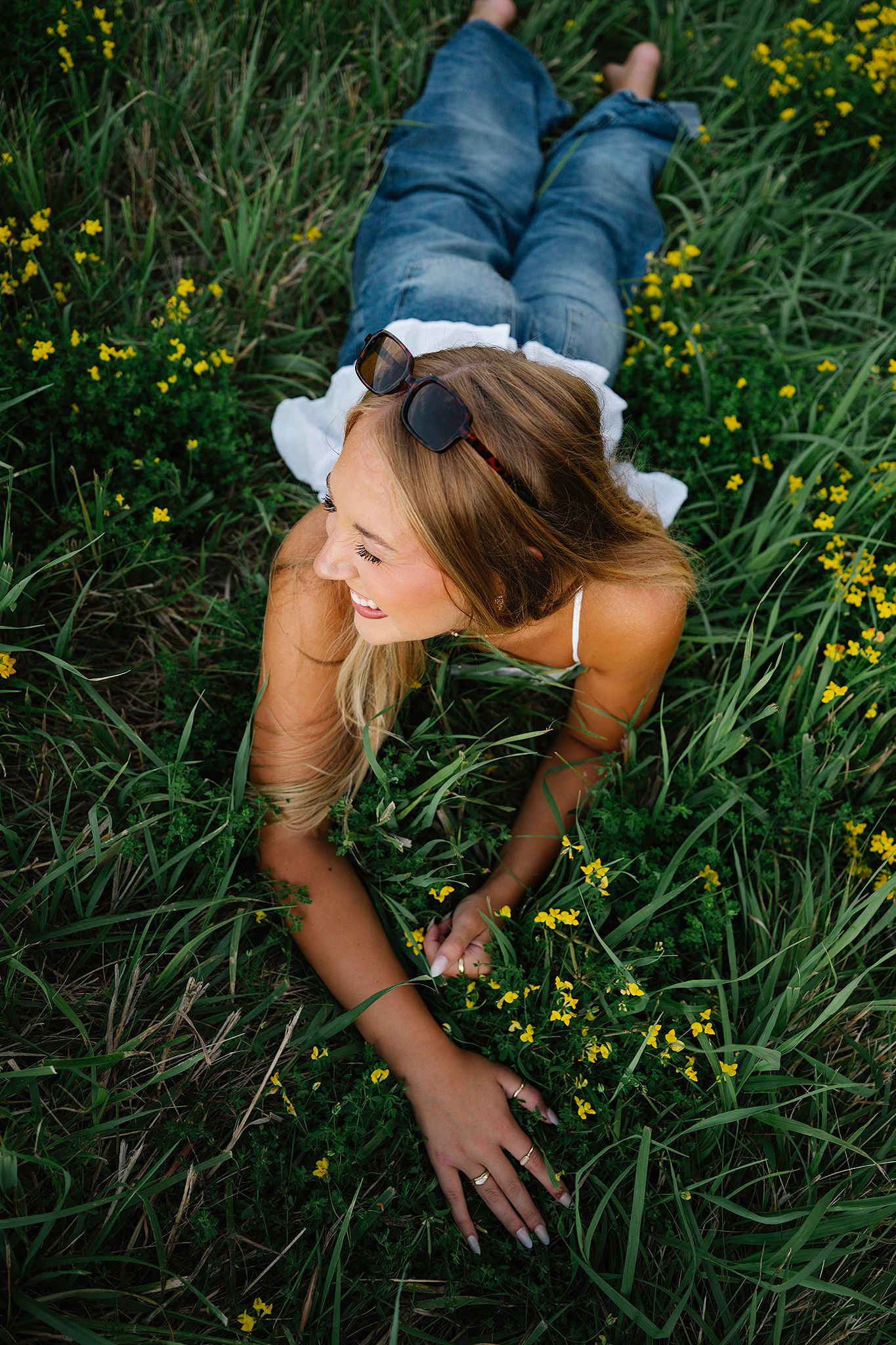 Young woman in jeans lying in green field with yellow wildflowers, smiling and touching blooms, sunglasses on head.