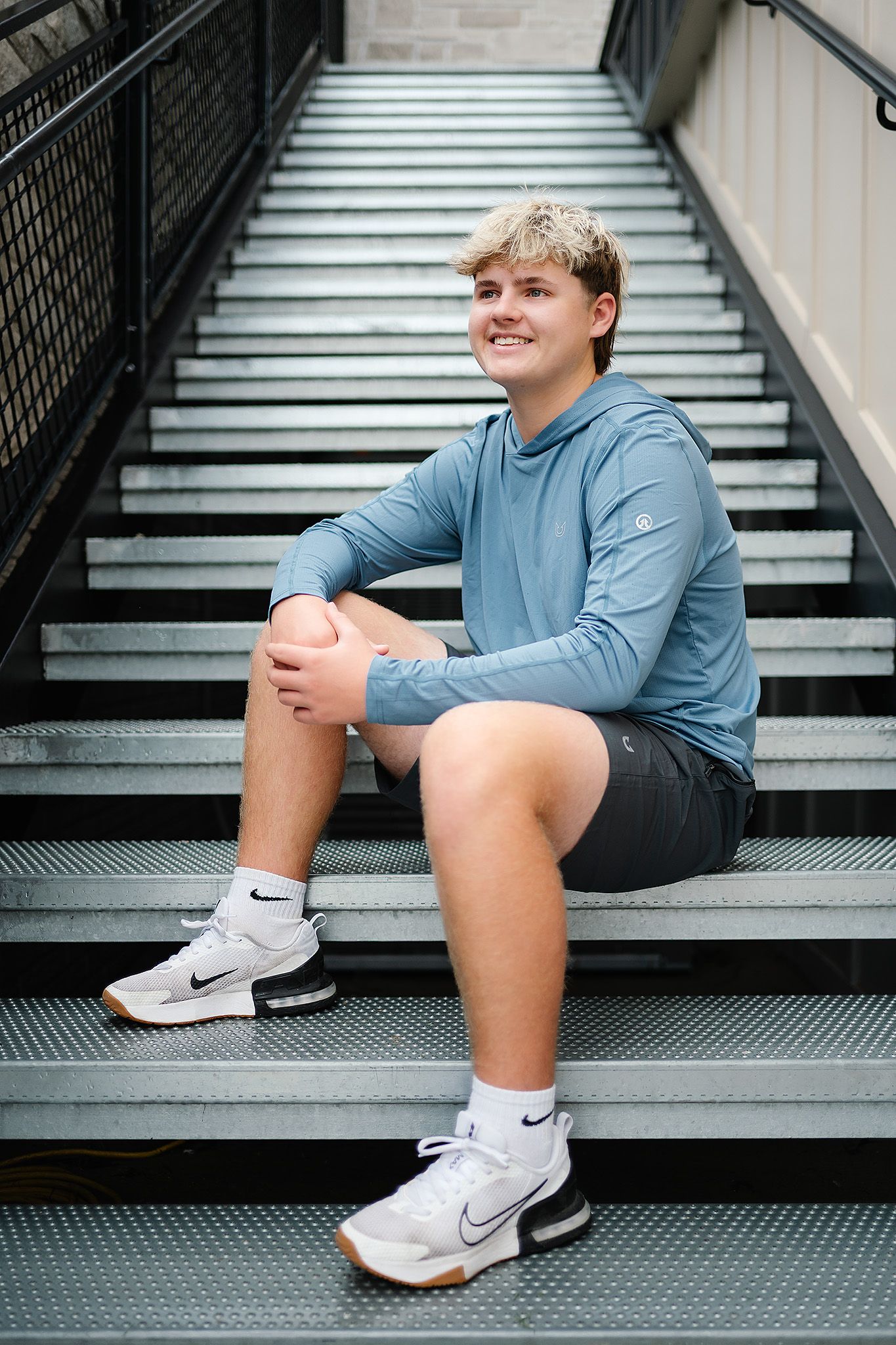 Young man in blue hoodie and Nike sneakers sitting on metal stairs, smiling outdoors.