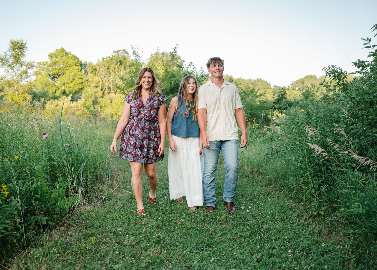 Three people smiling and walking together through a lush green meadow path during golden hour posing for Ankeny family photographer.