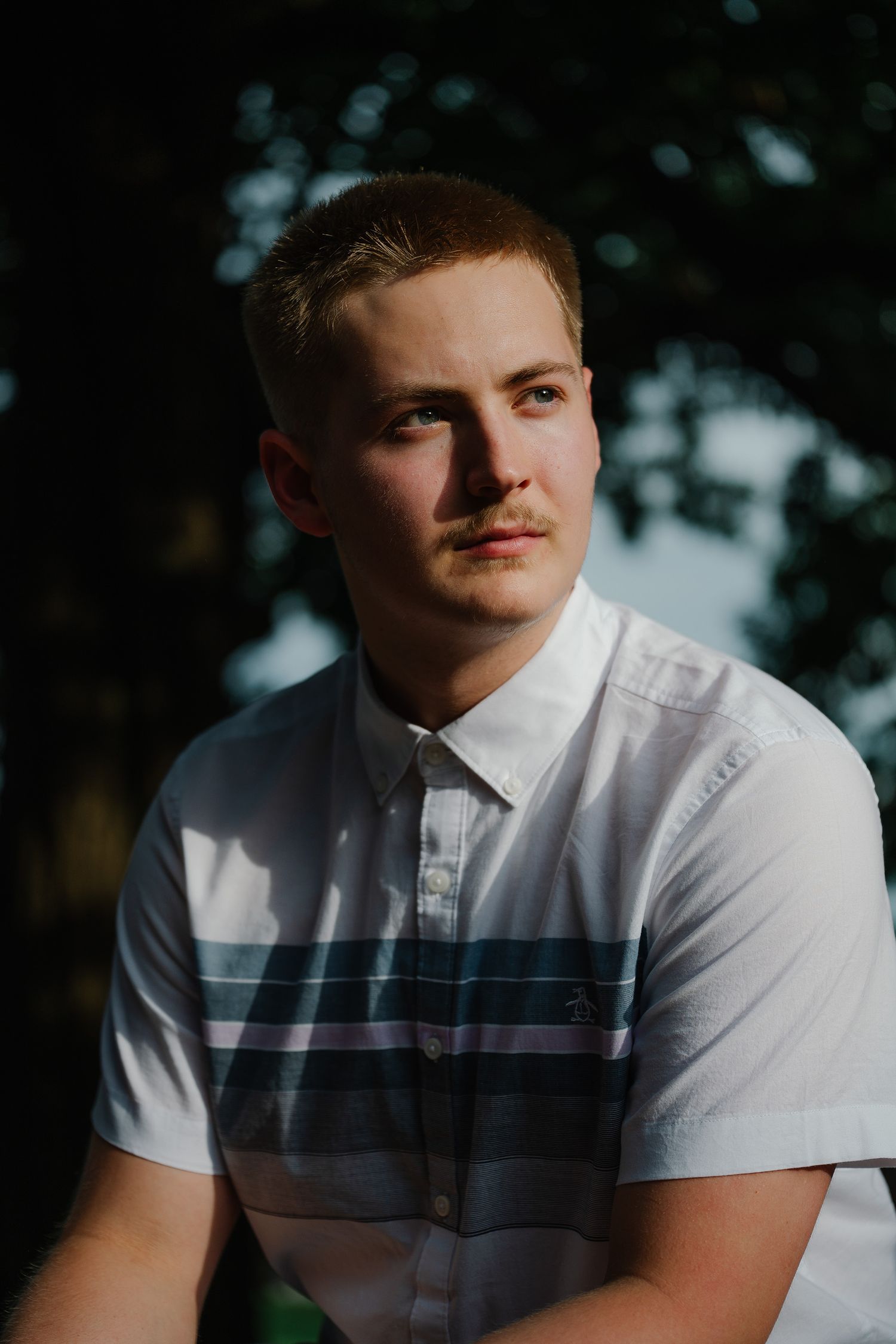 Young man in white striped polo shirt poses outdoors with soft golden light and green trees in background.