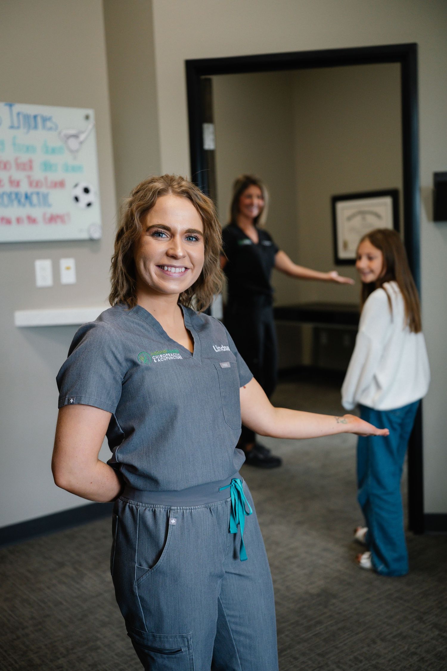 Smiling female healthcare worker in gray scrubs welcomes patients at a chiropractic office with staff and child in background.