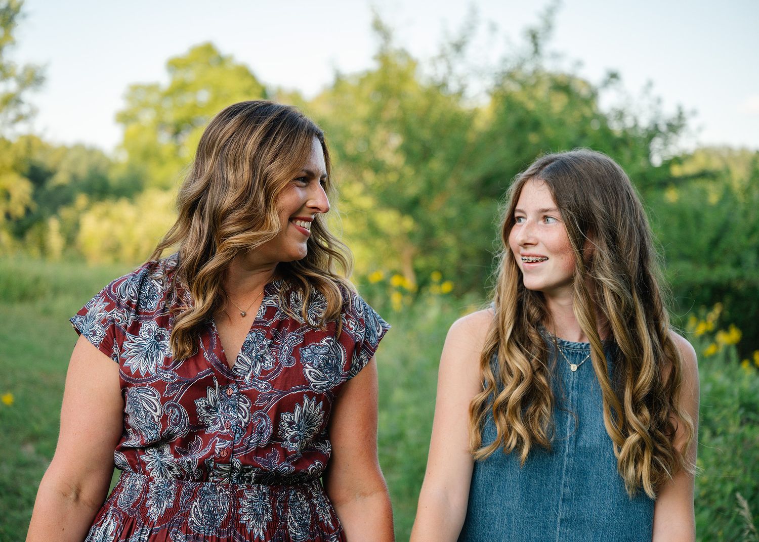 Two women smiling at each other outdoors in a green field, wearing floral and denim outfits posing for Ankeny family photographer.