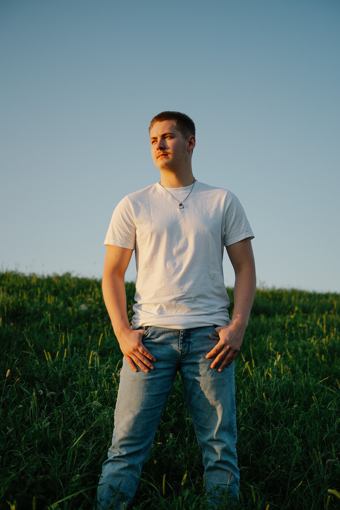 Young man in white t-shirt and jeans standing in green field under clear blue sky during golden hour.