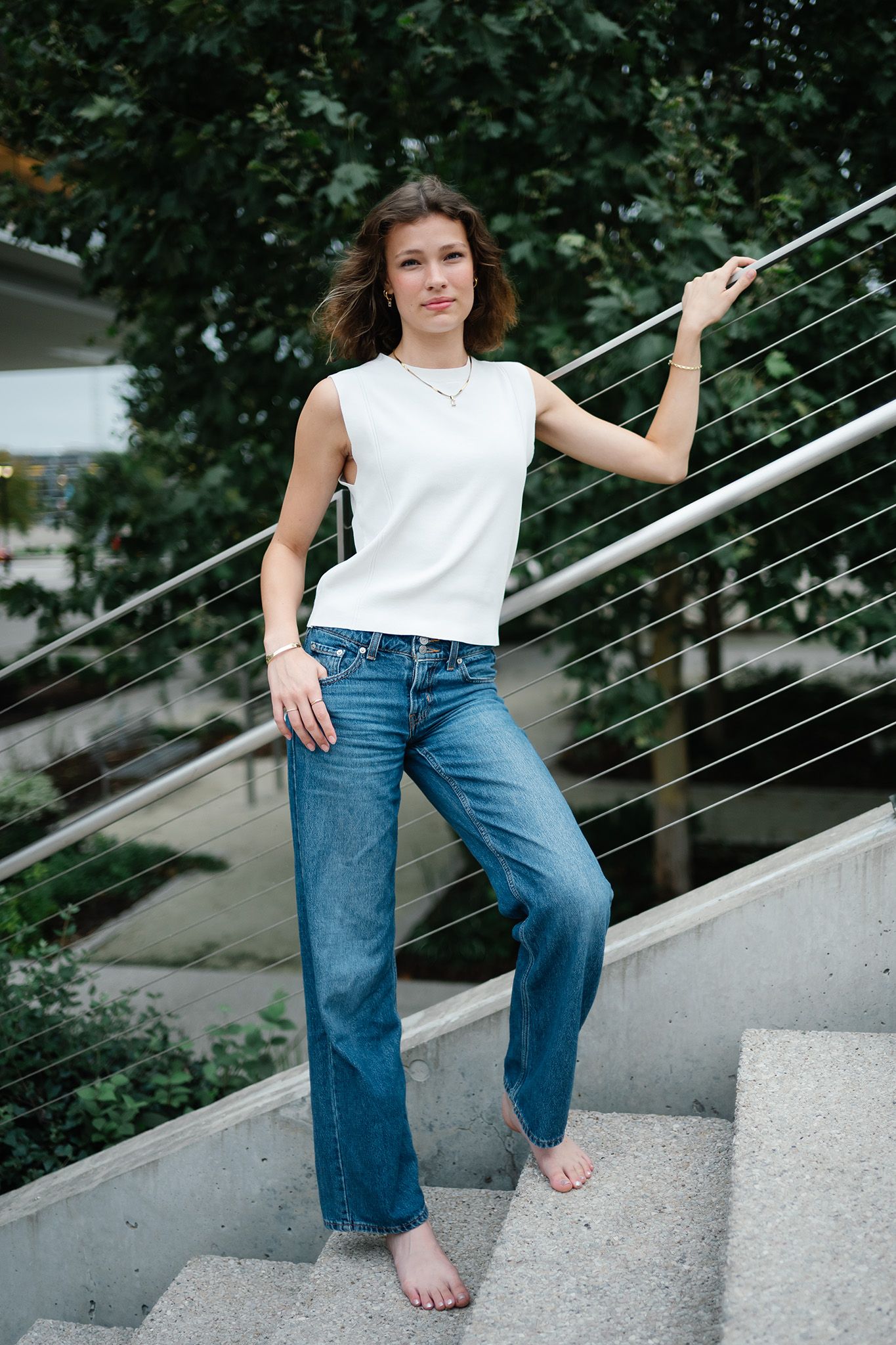 Young woman in white sleeveless top and wide-leg blue jeans standing barefoot on outdoor stairs.