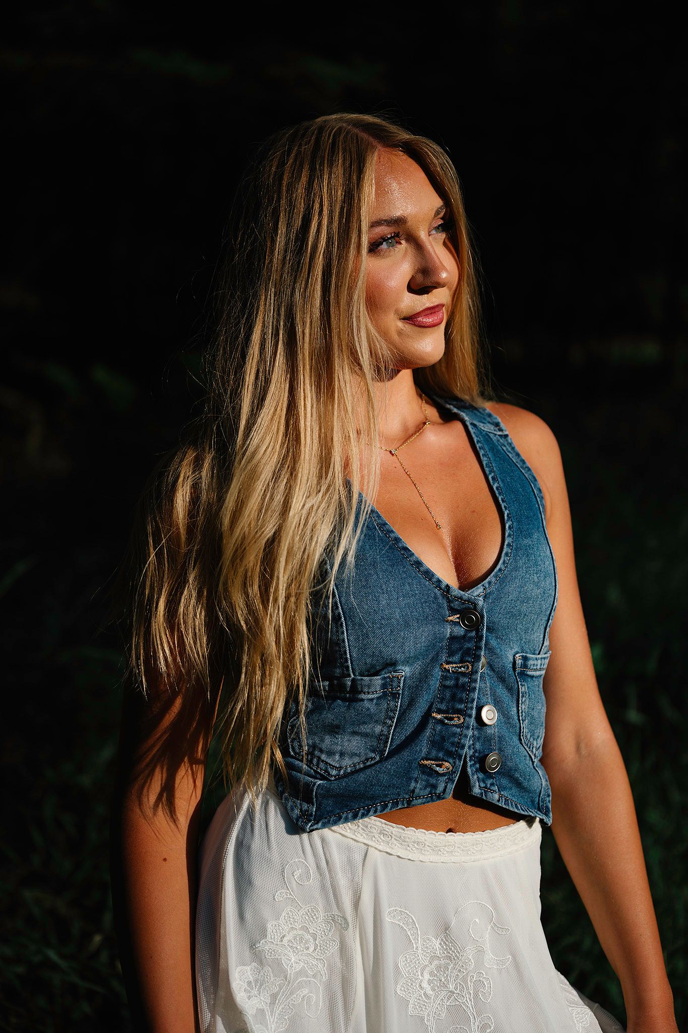 Young woman with long blonde hair wearing a denim vest and white skirt poses outdoors in golden hour light for Ankeny senior photographer