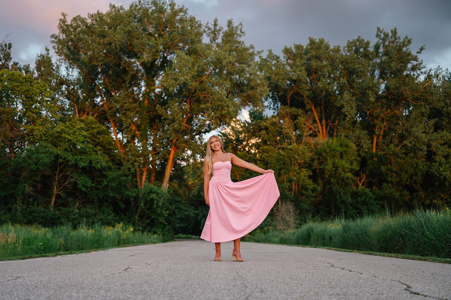 Woman in pink dress twirling on a path surrounded by lush green trees at sunset posing for Des Moines senior photographer