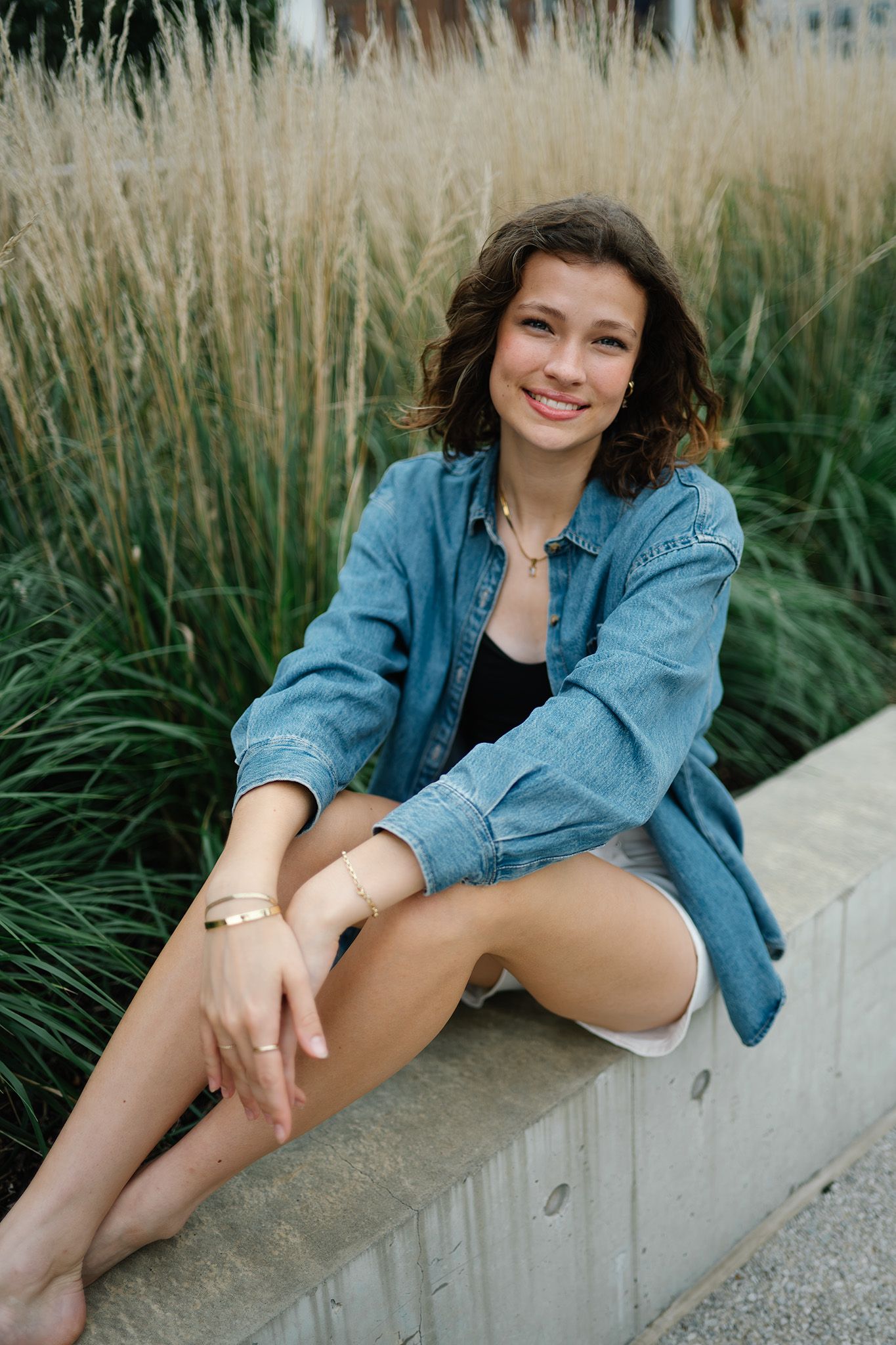 Young woman in denim shirt and white shorts sitting on concrete ledge, smiling outdoors near tall ornamental grasses.