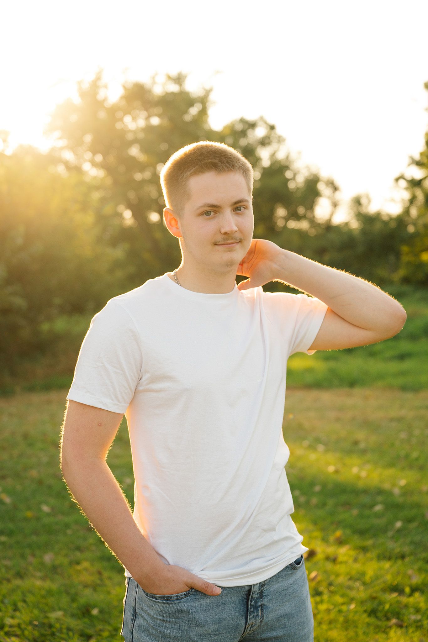 Young man in white t-shirt and jeans posing outdoors in golden sunset light with green trees in background.
