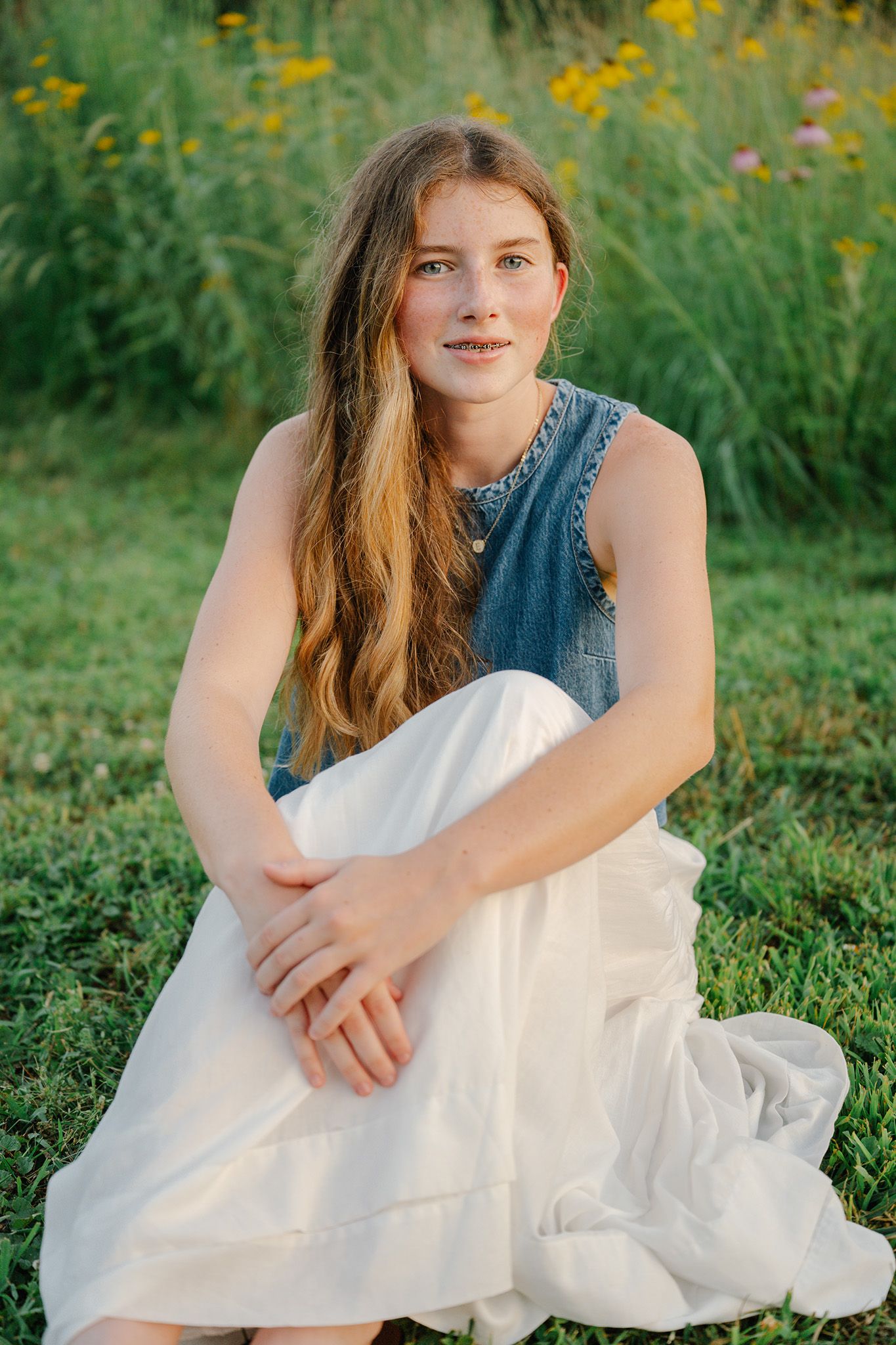 Young woman with long hair sits on grass in a denim vest and white skirt, by wildflowers for Ankeny senior photographer.
