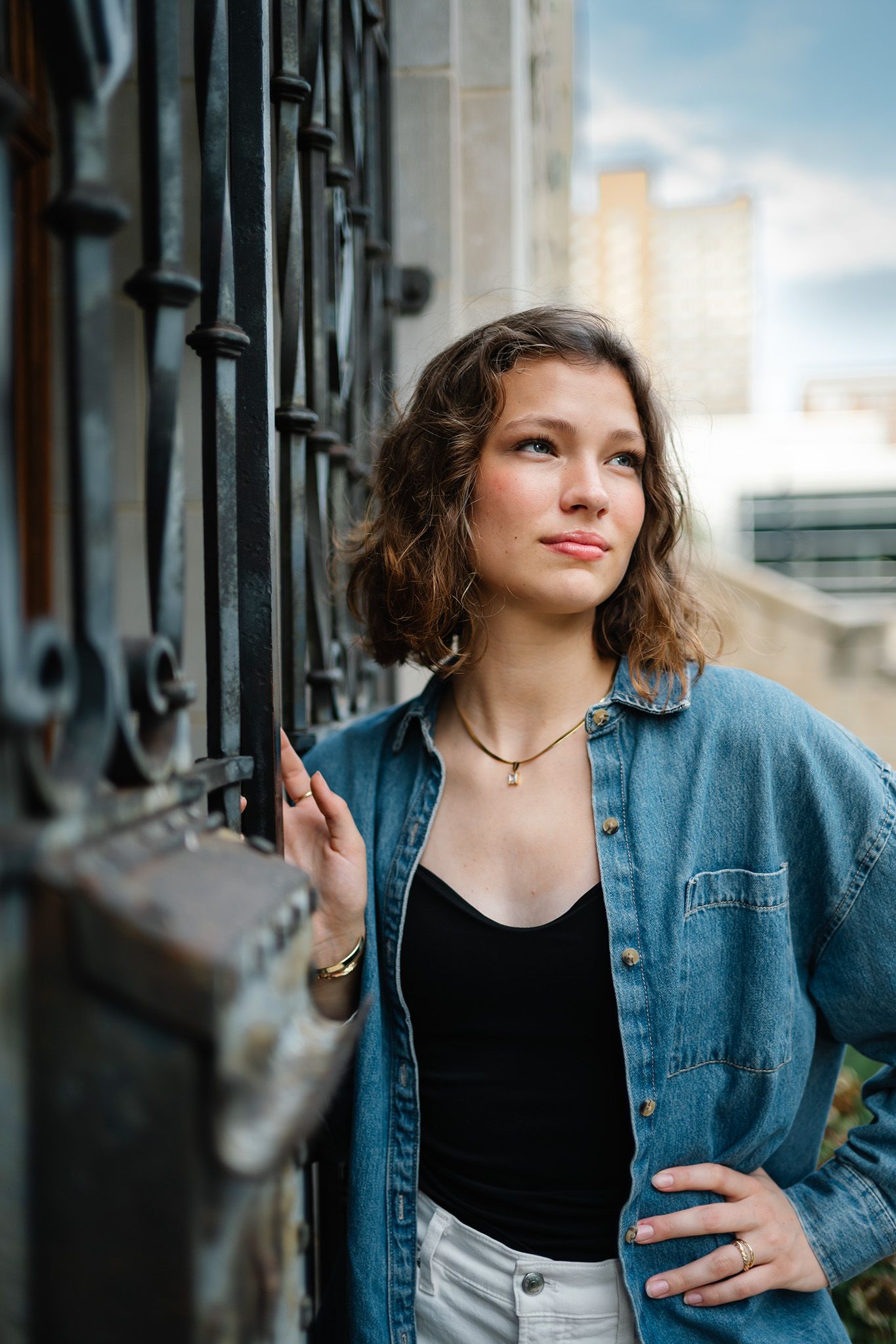 Young woman in denim shirt and black top leans against ornate iron gate, gazing upward in urban setting.