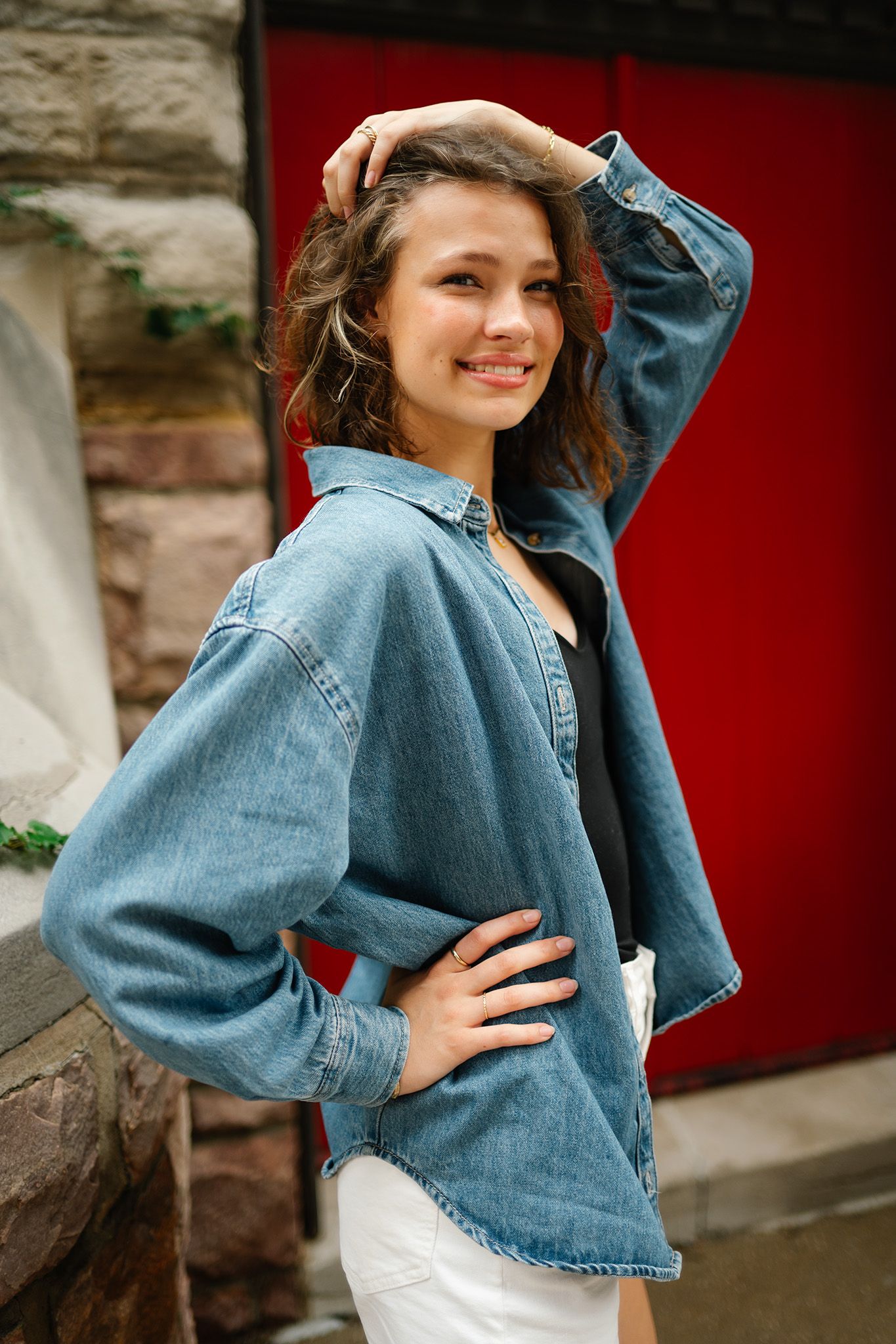 Young woman in oversized denim shirt and white skirt posing outdoors near red door, smiling confidently.