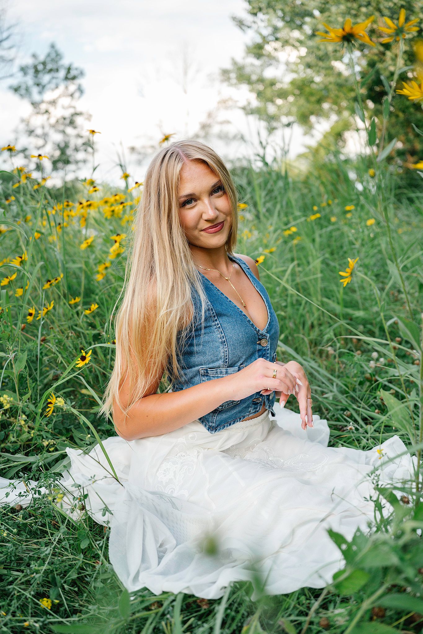 Blonde woman in denim vest and white skirt sitting among yellow wildflowers in a lush green field posing for Des Moines senior photographer