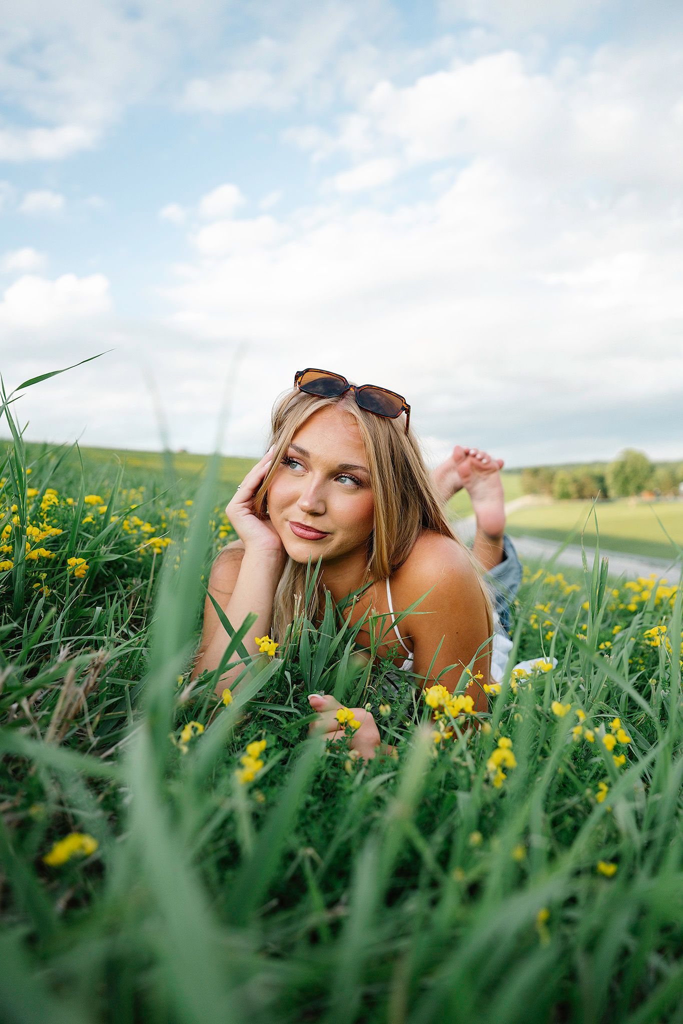 Blonde woman lying in green field with yellow wildflowers, sunglasses on head, gazing thoughtfully for Ankeny senior photographer