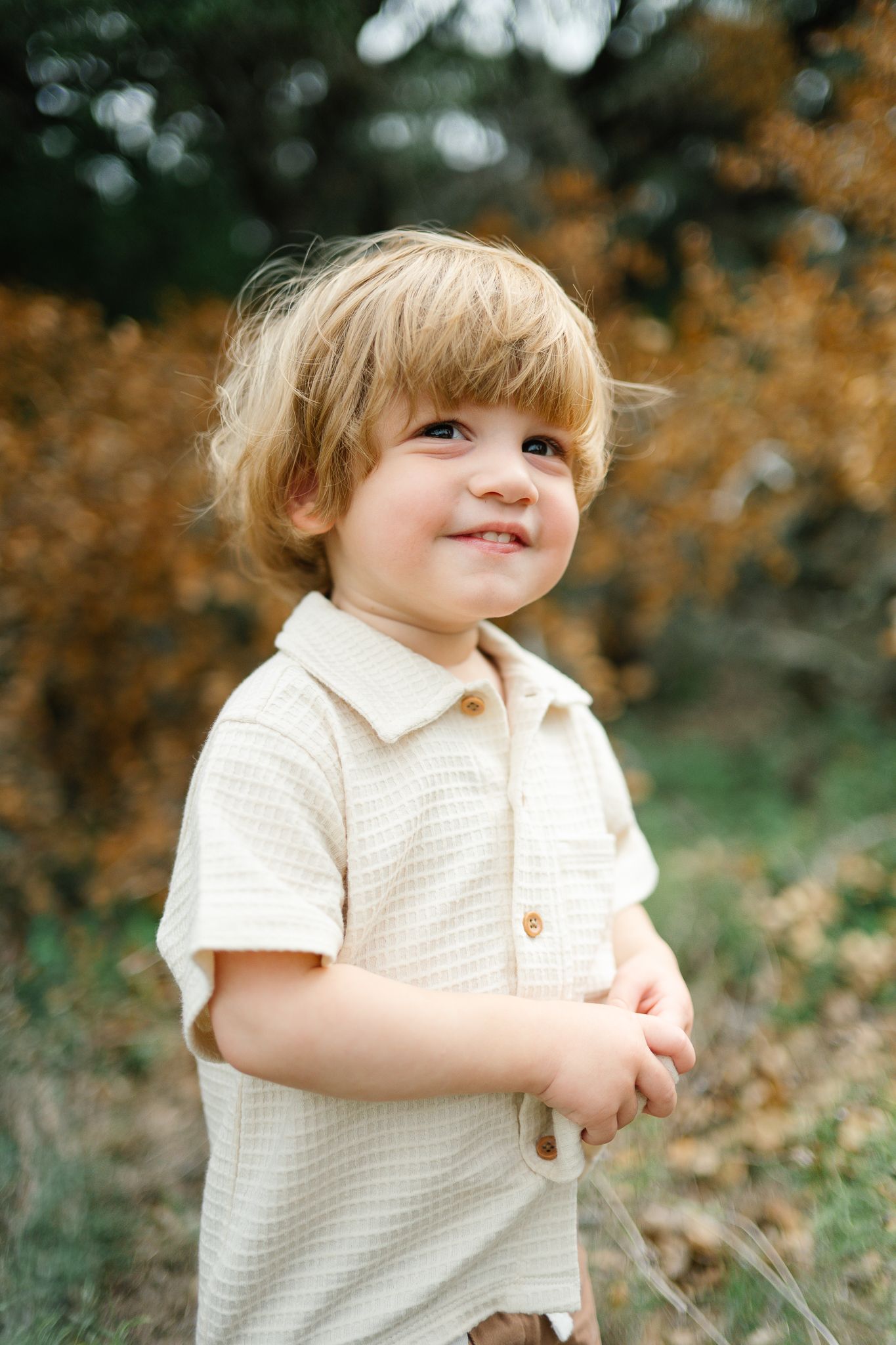 Smiling toddler with tousled blond hair in a cream striped collared shirt stands outdoors among golden autumn foliage.